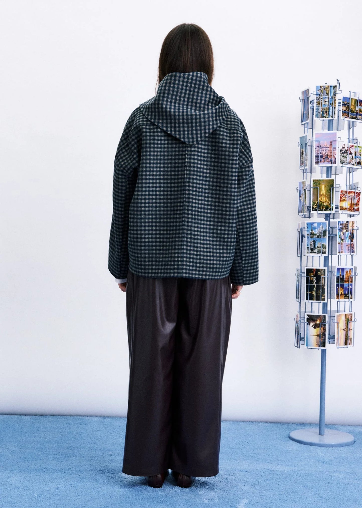 A person stands with their back to the camera wearing a dark green and black gingham patterned hooded jacket and wide-leg dark brown pants, standing in a room with a light blue floor against a white wall next to a clothing rack.