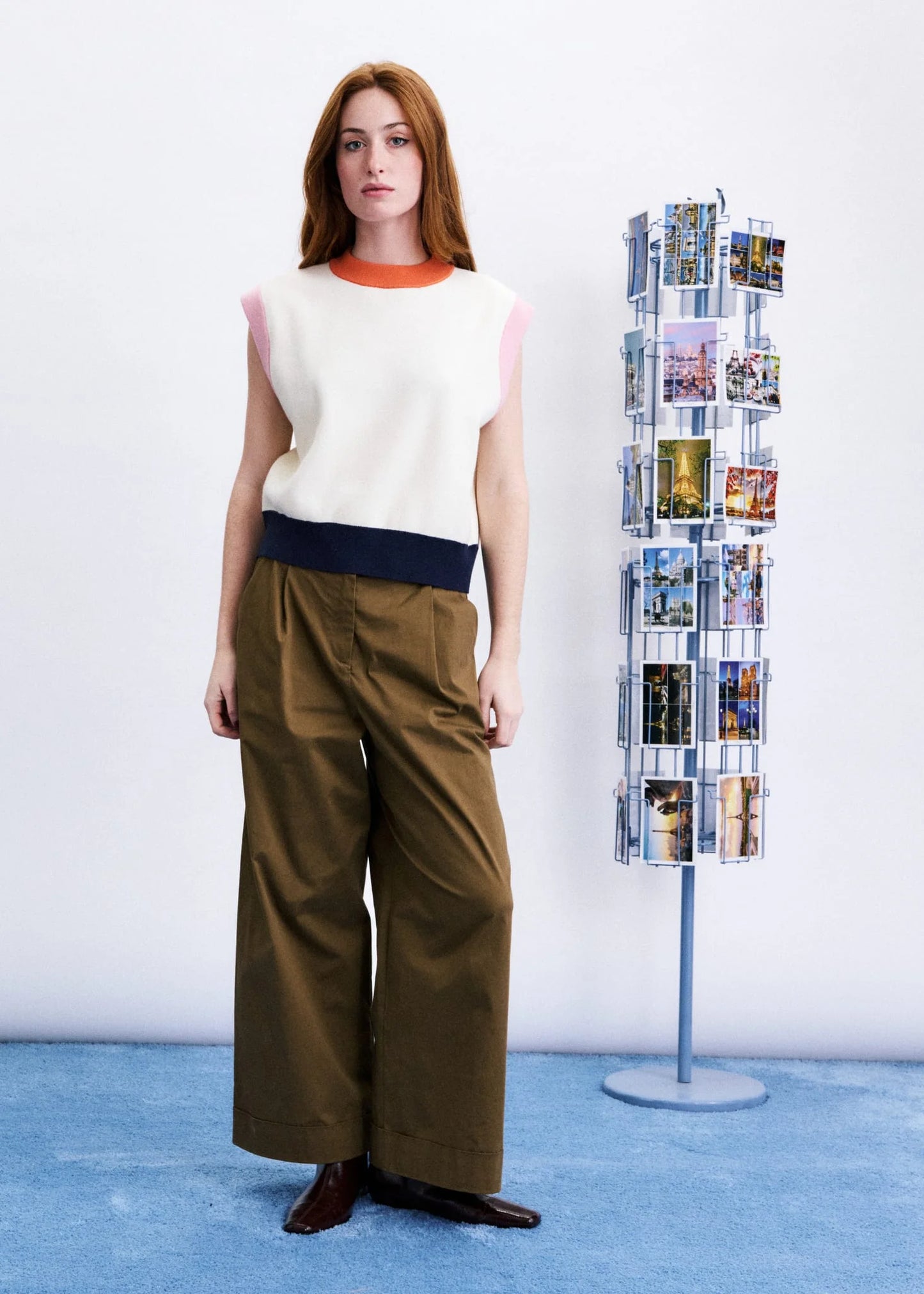 A woman stands in a studio wearing a sleeveless knit top with cream, pink, and navy color-blocked panels, paired with wide-leg brown pants. A tall rotating photo display stand is visible to her right against a white wall and blue floor.
