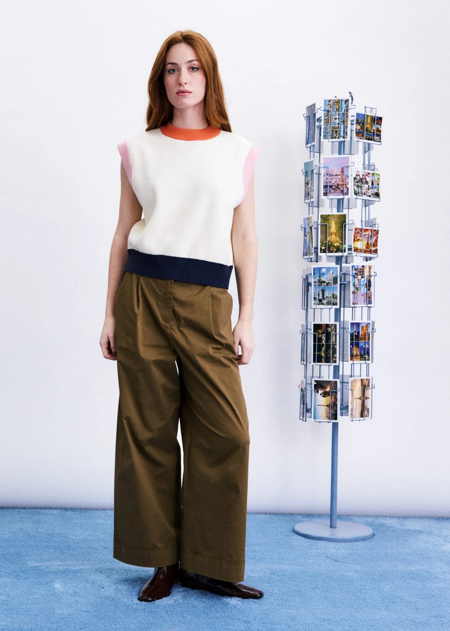 A woman stands in a studio wearing a sleeveless knit top with cream, pink, and navy color-blocked panels, paired with wide-leg brown pants. A tall rotating photo display stand is visible to her right against a white wall and blue floor.