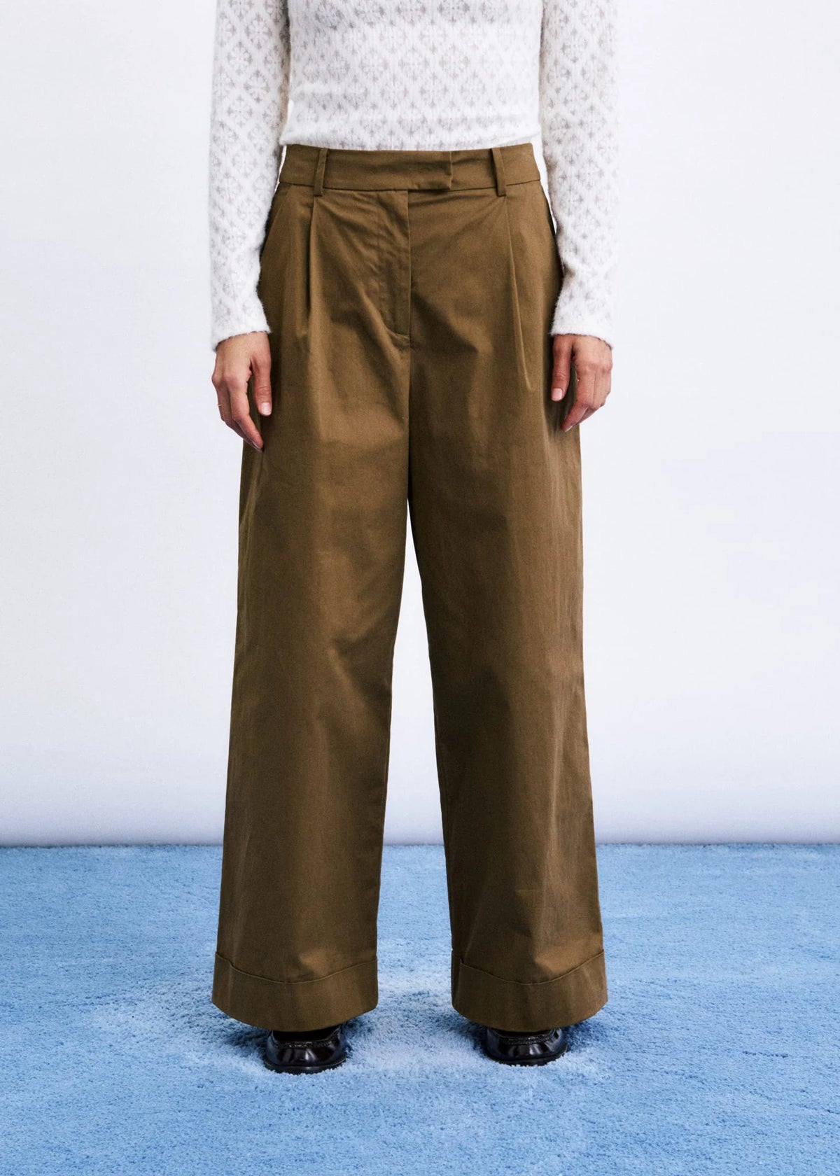 A model is shown from the chest down wearing high-waisted, wide-leg brown trousers with pleats, paired with a white patterned long-sleeve top and black shoes, standing on a blue textured floor against a white wall.