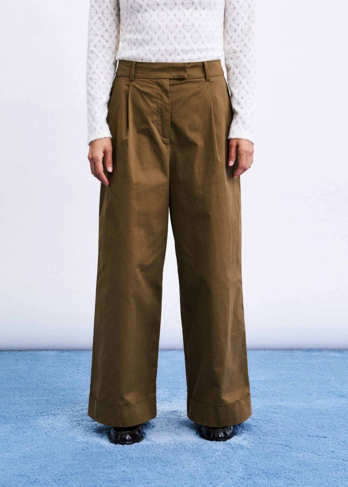 A model is shown from the chest down wearing high-waisted, wide-leg brown trousers with pleats, paired with a white patterned long-sleeve top and black shoes, standing on a blue textured floor against a white wall.