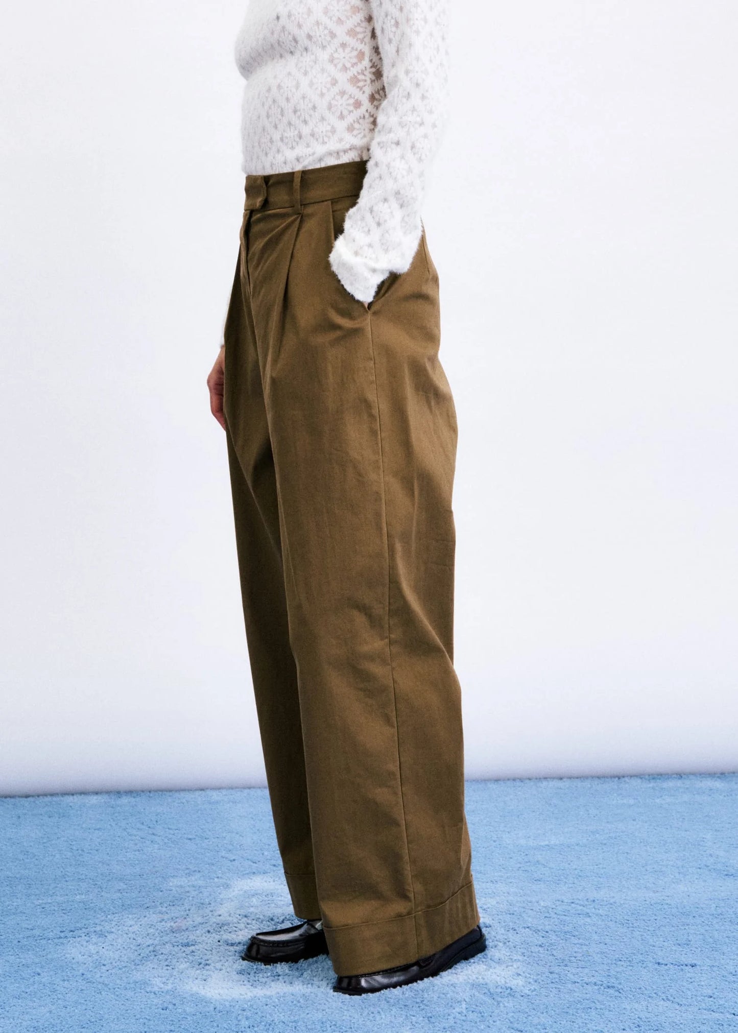 A person wearing high-waisted, wide-leg brown trousers with a white textured knit top and black shoes, standing against a white wall on a blue floor.