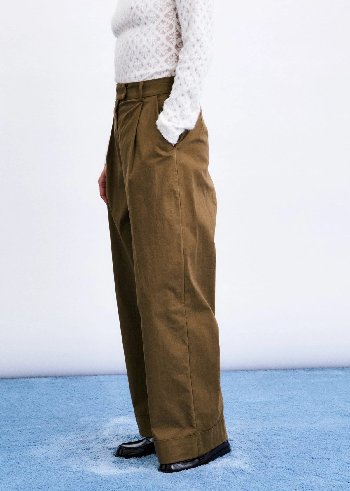 A person wearing high-waisted, wide-leg brown trousers with a white textured knit top and black shoes, standing against a white wall on a blue floor.