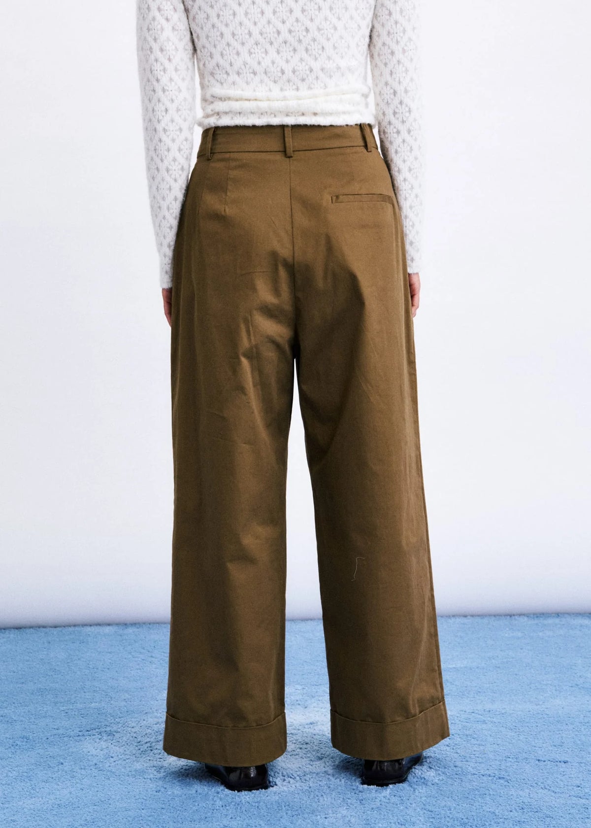 A person shown from the back wearing wide-leg brown trousers and a white textured knit top, standing against a white wall with a blue floor.