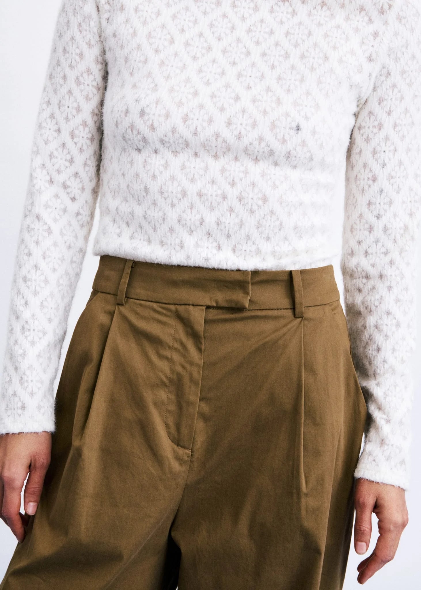 A close-up of a person wearing a white textured knit sweater and olive brown pleated trousers, cropped from the neck down against a plain light background.