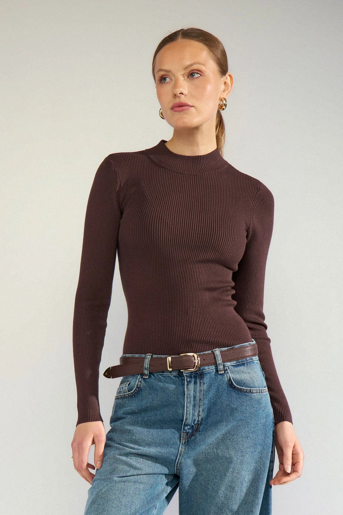 A woman wearing a dark brown ribbed mock neck long-sleeve top and blue jeans with a brown belt, standing against a light gray studio background.
