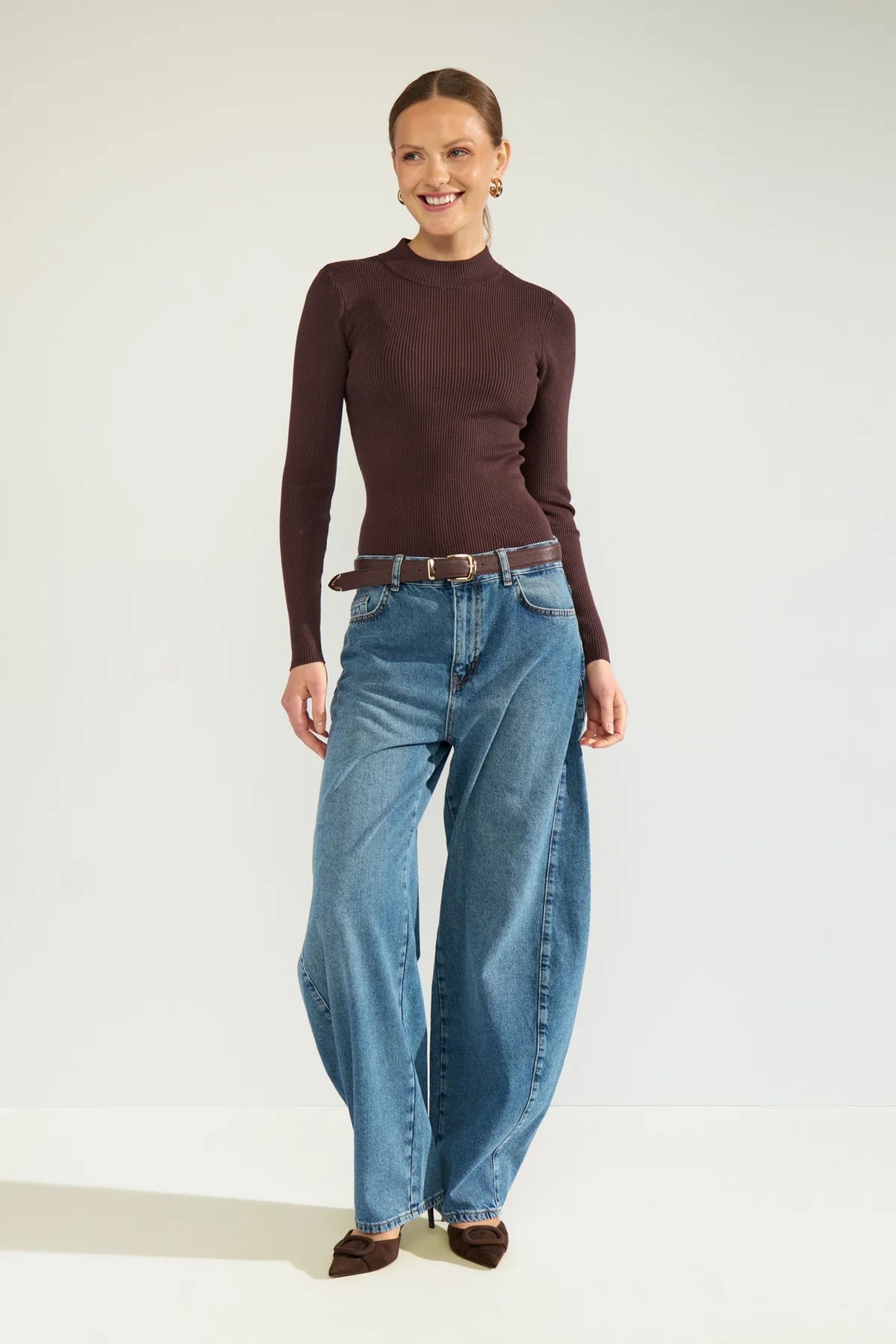 A woman modeling a dark brown mock-neck long-sleeve top and wide-leg blue jeans with a brown belt, standing against a plain light gray studio background.