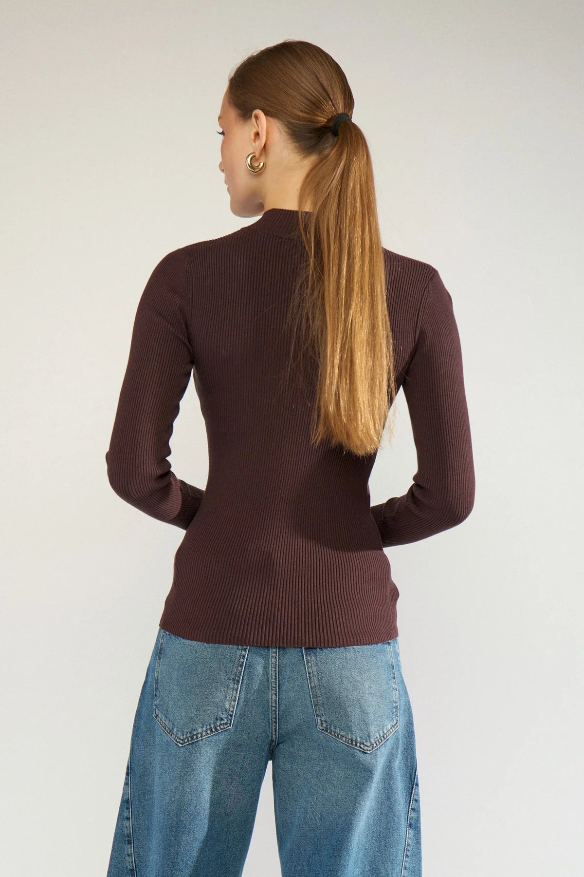 A woman shown from the back wearing a fitted ribbed long-sleeve mock-neck sweater in dark plum brown, paired with wide-leg blue jeans, against a plain light gray studio background.