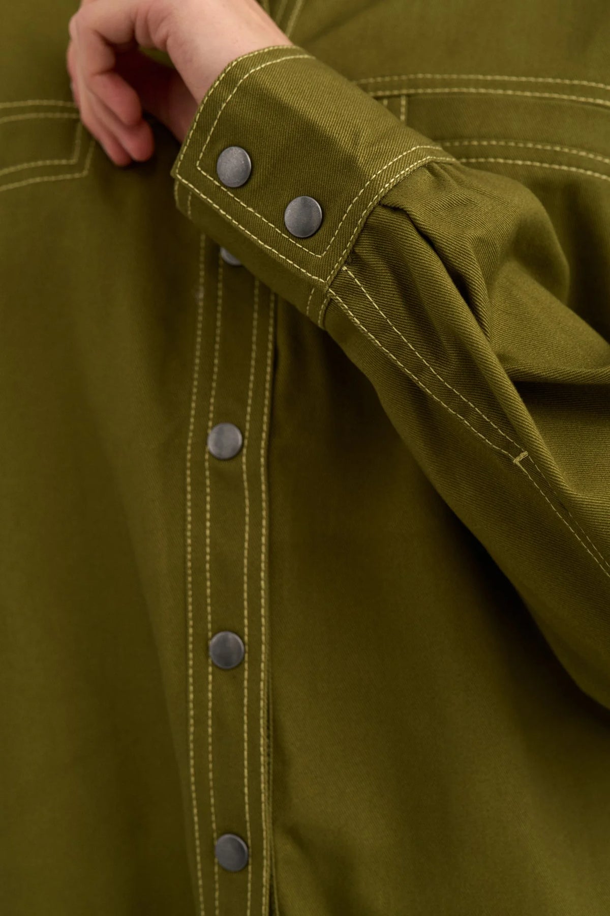Close-up of an olive green button-front garment with visible contrast stitching and dark metal buttons, including a cuff detail with three buttons and a hand adjusting the sleeve.