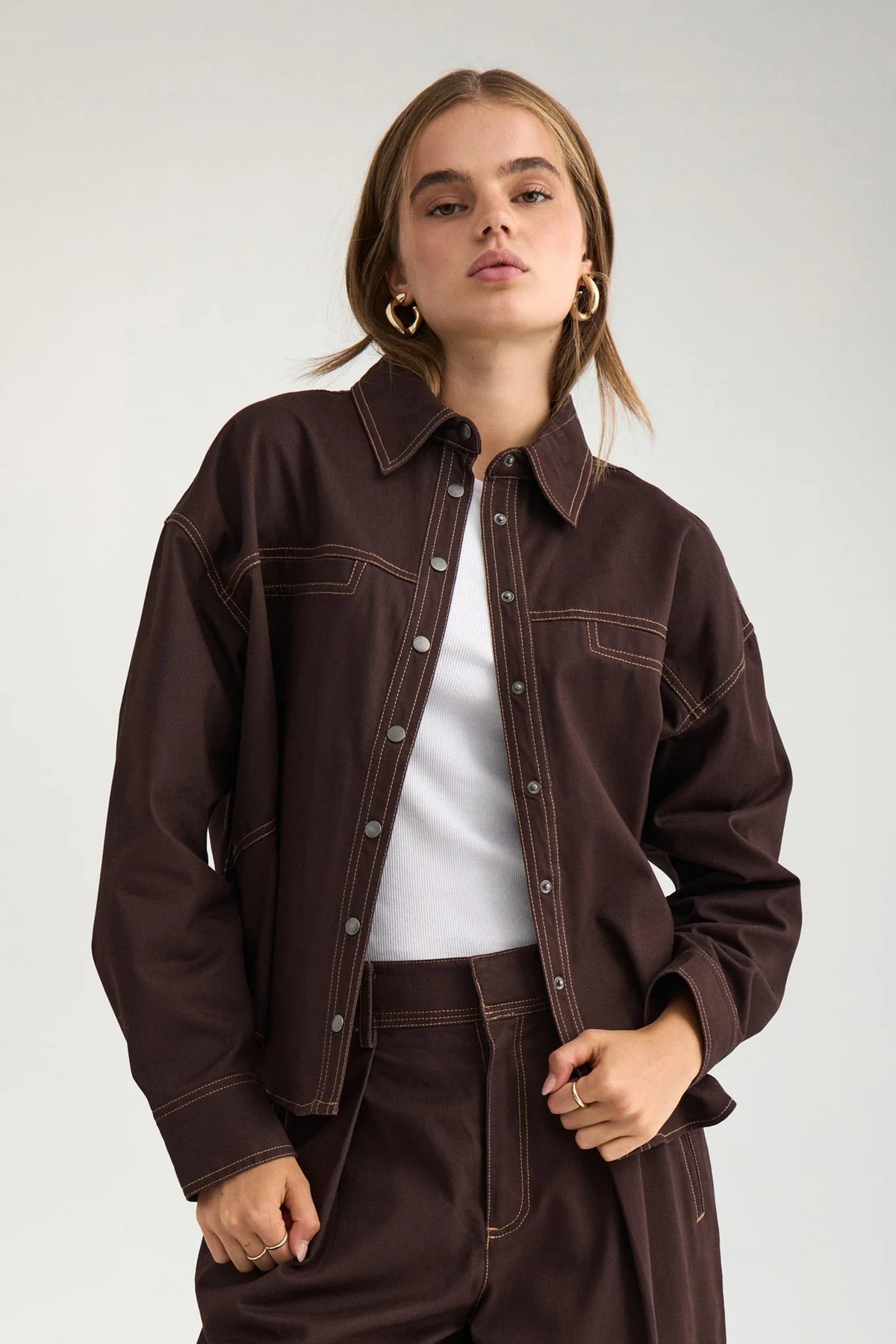 A model wearing a dark brown cropped denim-style jacket with contrast stitching over a white top and matching dark brown trousers, set against a plain light gray studio background.