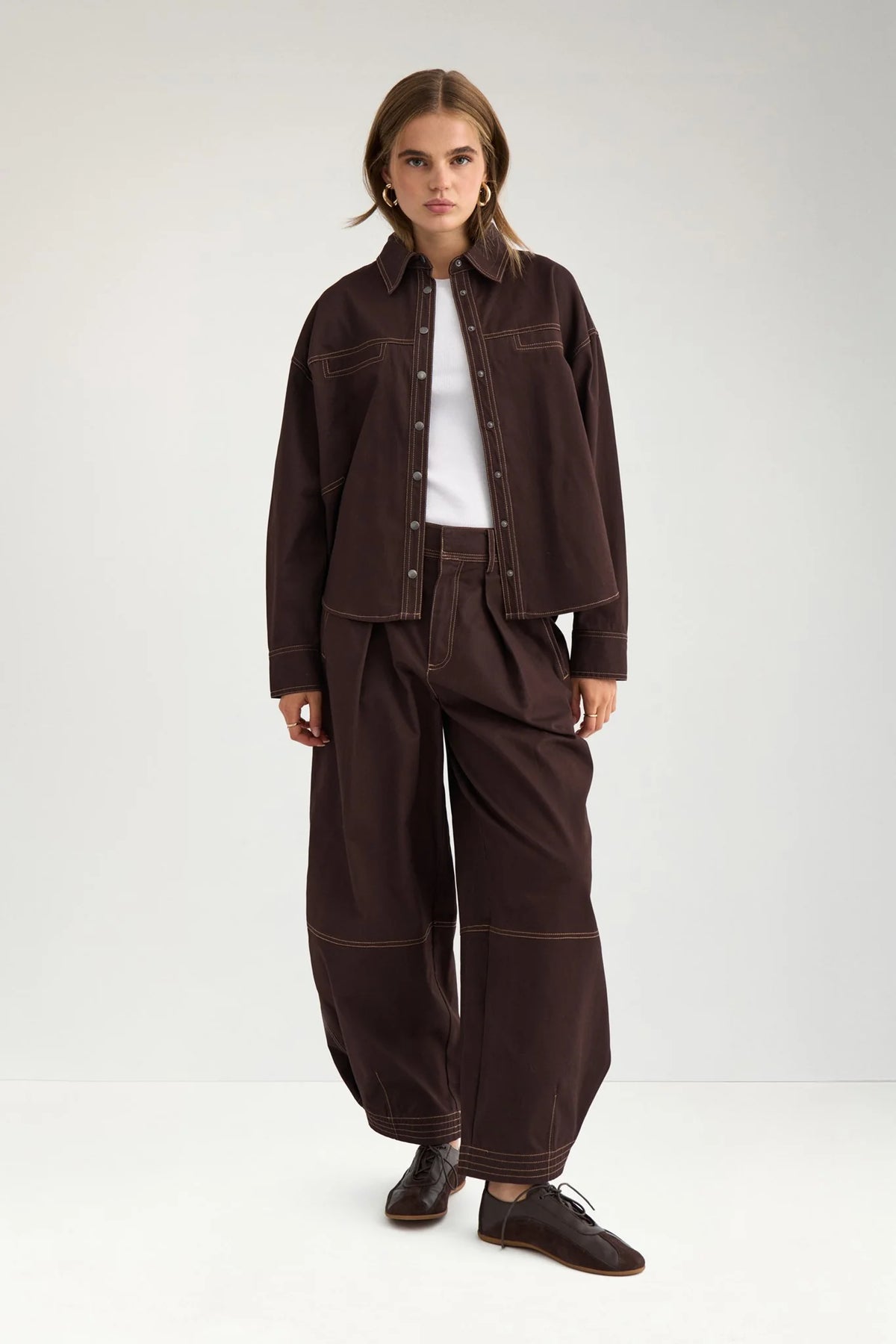 A model stands against a light gray studio background wearing a dark brown cropped button-up shirt jacket, matching wide-leg pants, a white top, and dark brown shoes.