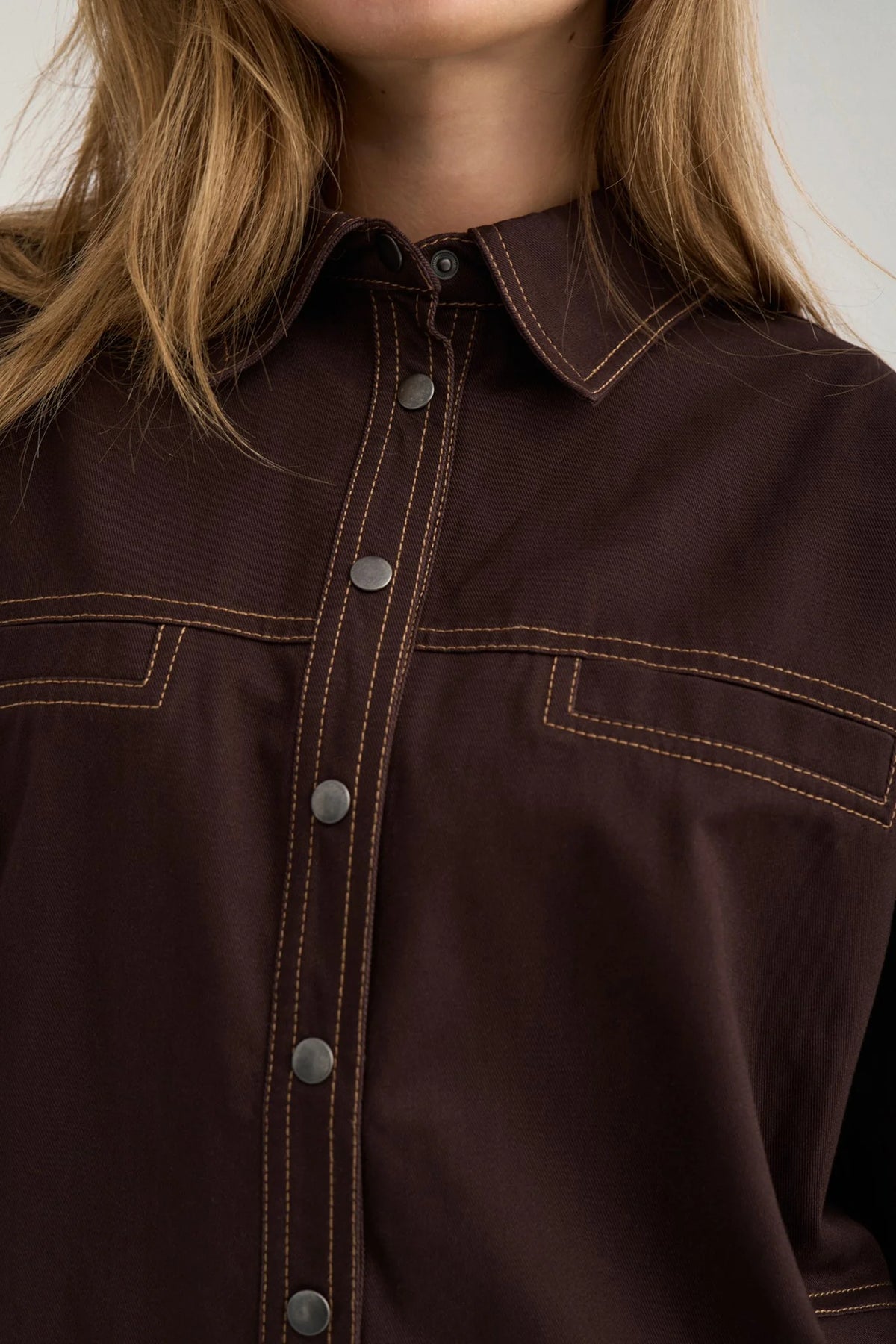 Close-up of a person wearing a dark brown button-front shirt with contrast tan stitching and silver buttons.