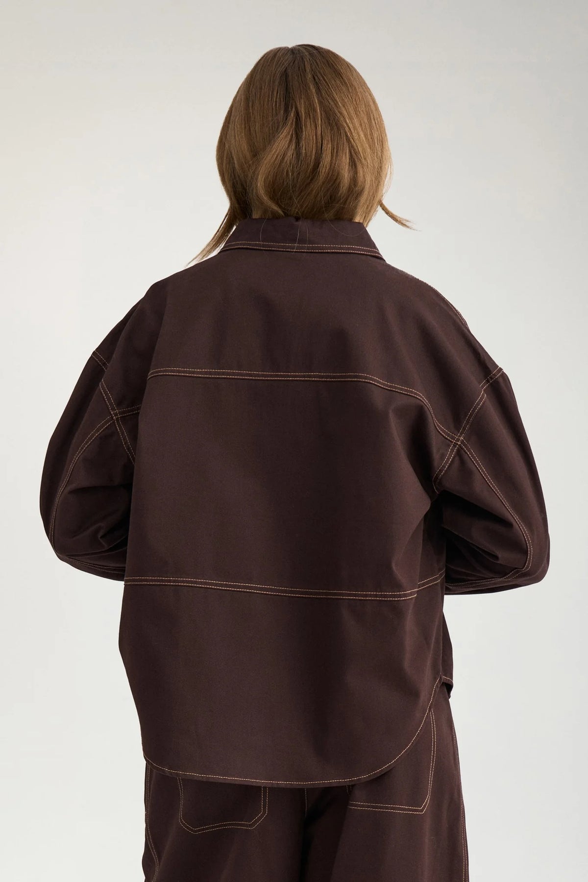 A person shown from the back wearing a dark brown oversized jacket with contrasting light brown stitching, paired with matching brown pants, against a plain light gray studio background.
