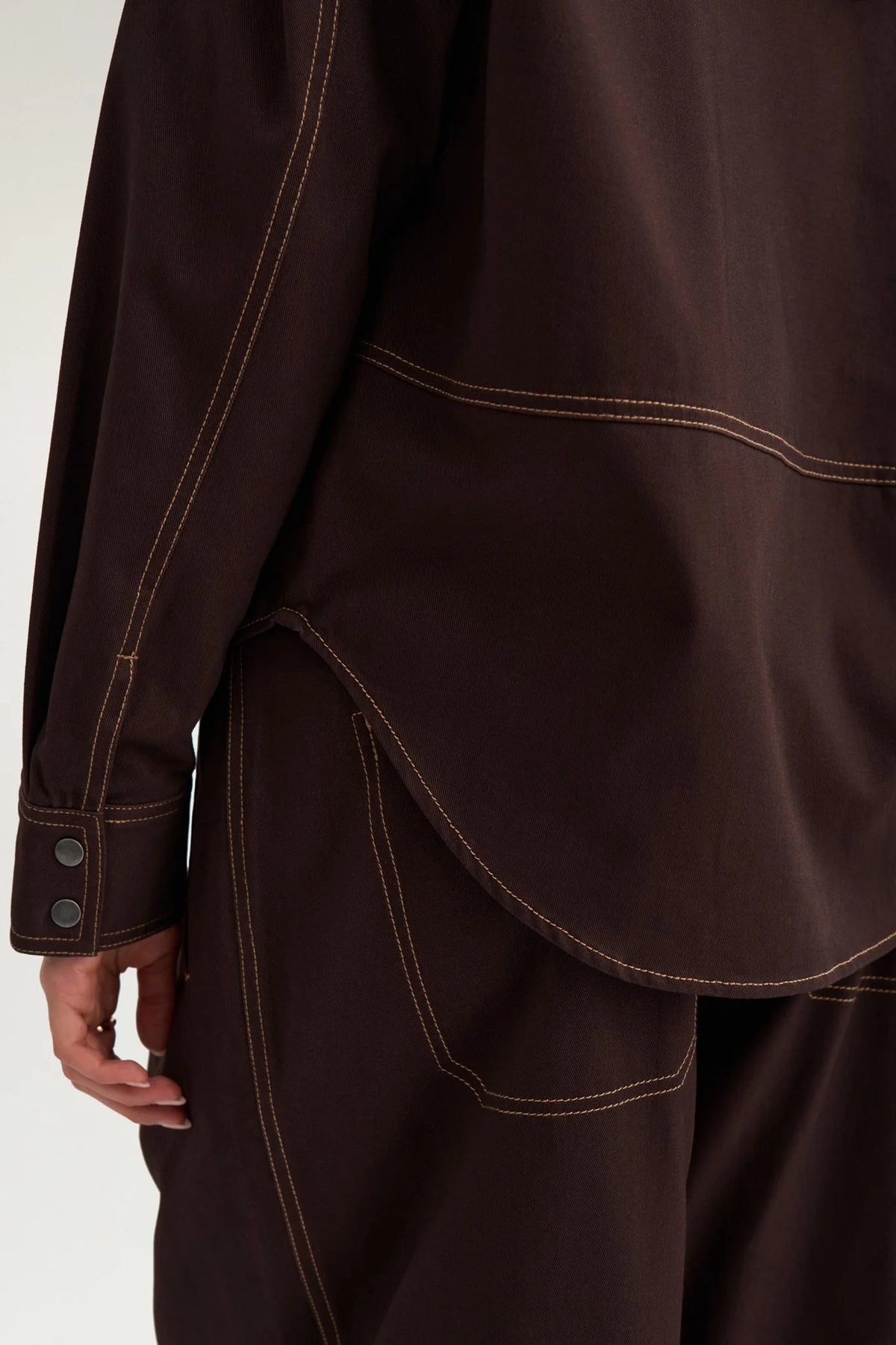 Close-up of a dark brown jacket or overshirt with contrasting light brown stitching, featuring a buttoned cuff and a curved hem, worn by a model.