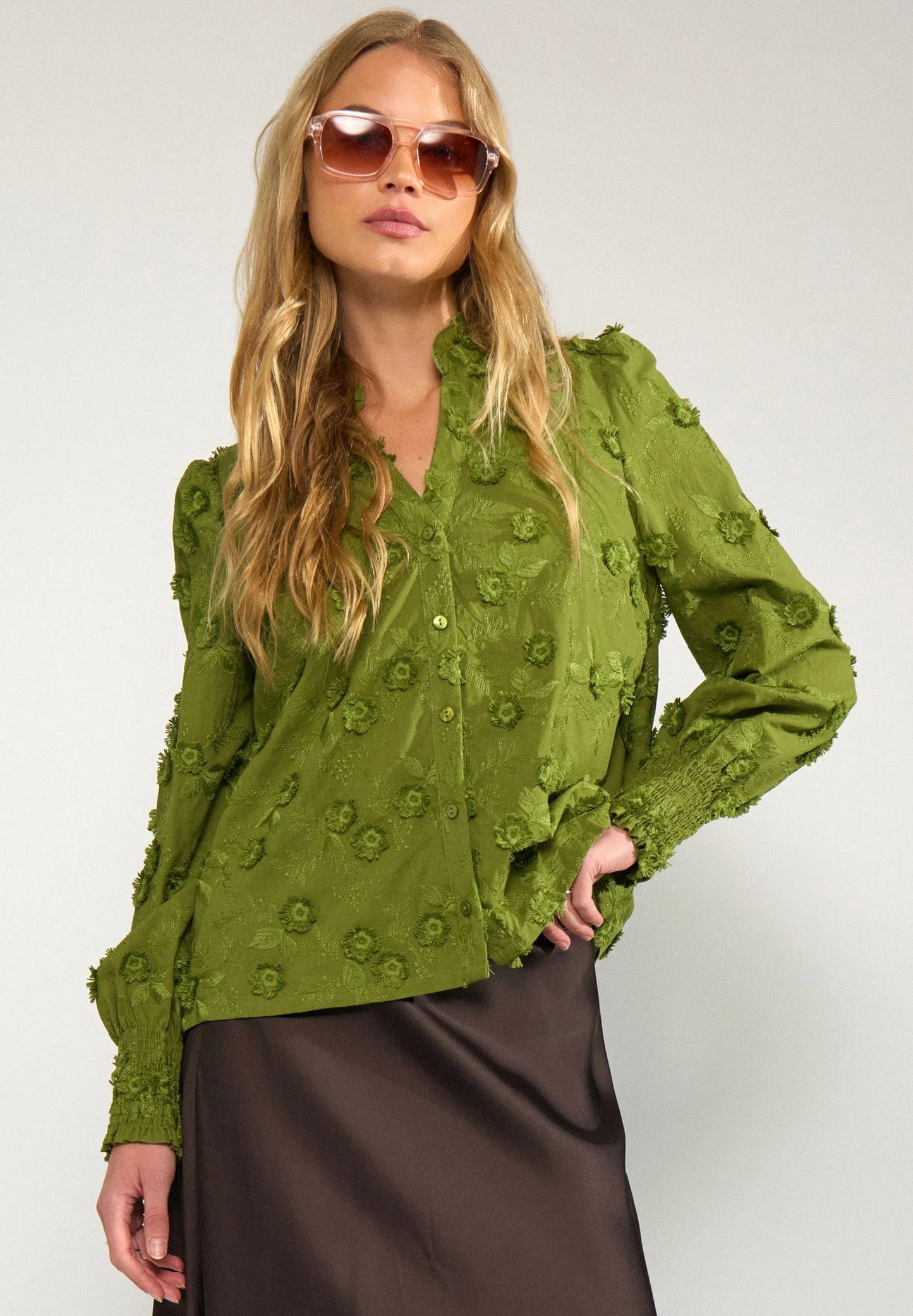 A woman modeling a textured olive green long-sleeve blouse with 3D floral embroidery and ruffled trim, paired with a dark brown satin skirt and sunglasses, against a light gray studio background.