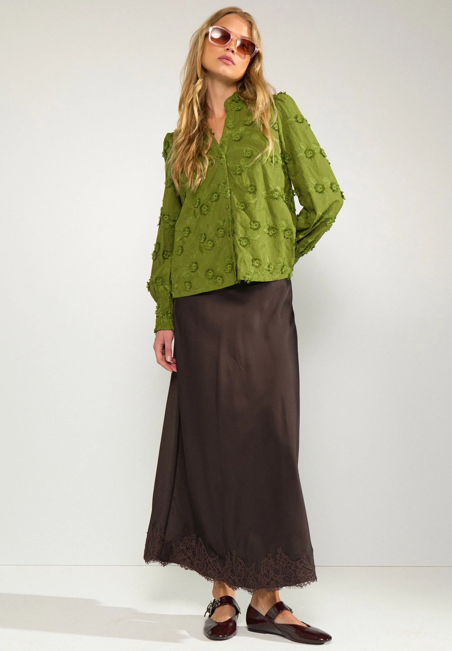 A woman modeling a green textured long-sleeve blouse and a dark brown satin maxi skirt with lace trim, paired with brown flat shoes against a plain light gray studio background.
