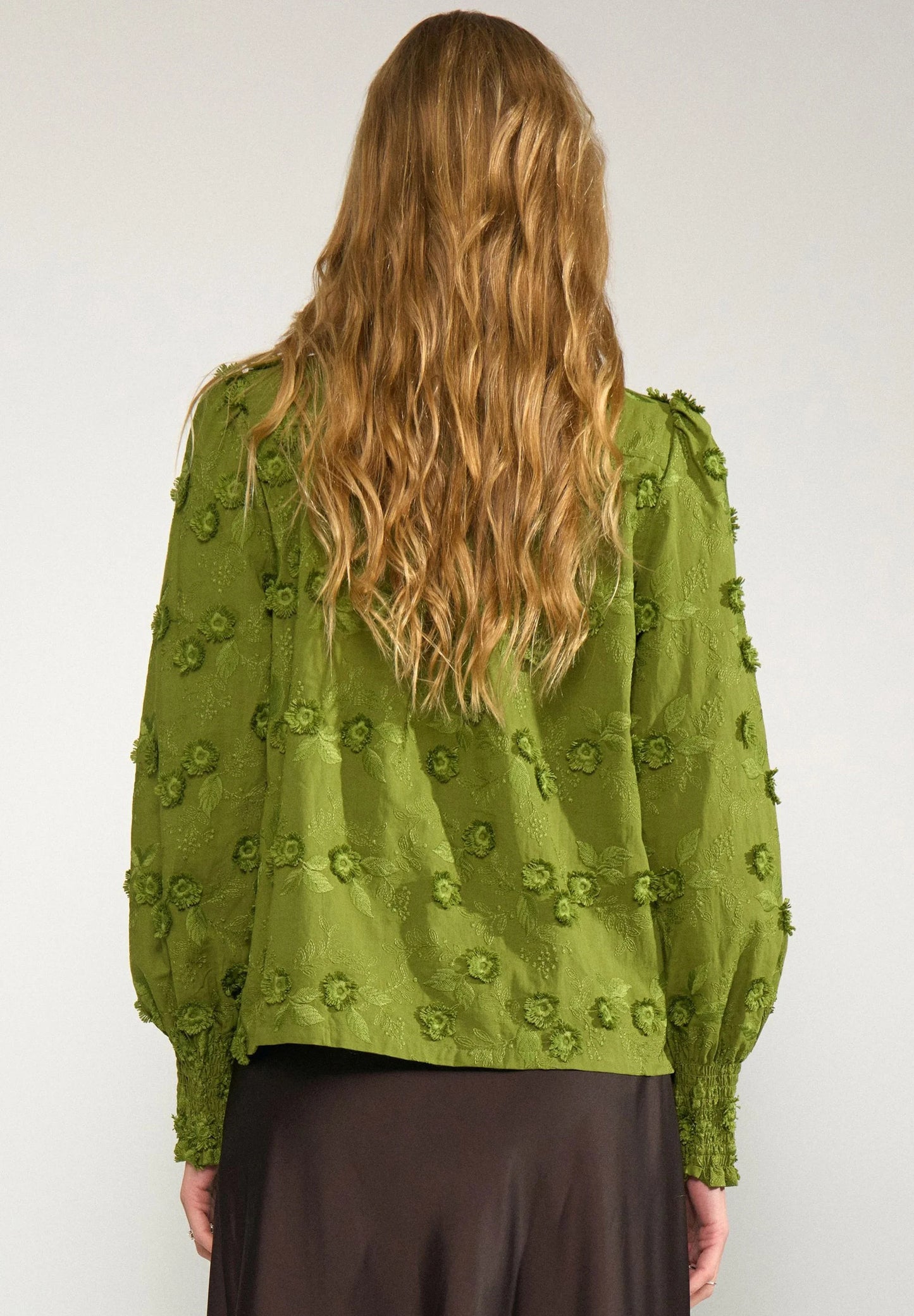 A woman viewed from the back wearing a textured olive green blouse with 3D floral embroidery and long puff sleeves, paired with a dark brown skirt, against a plain light gray background.