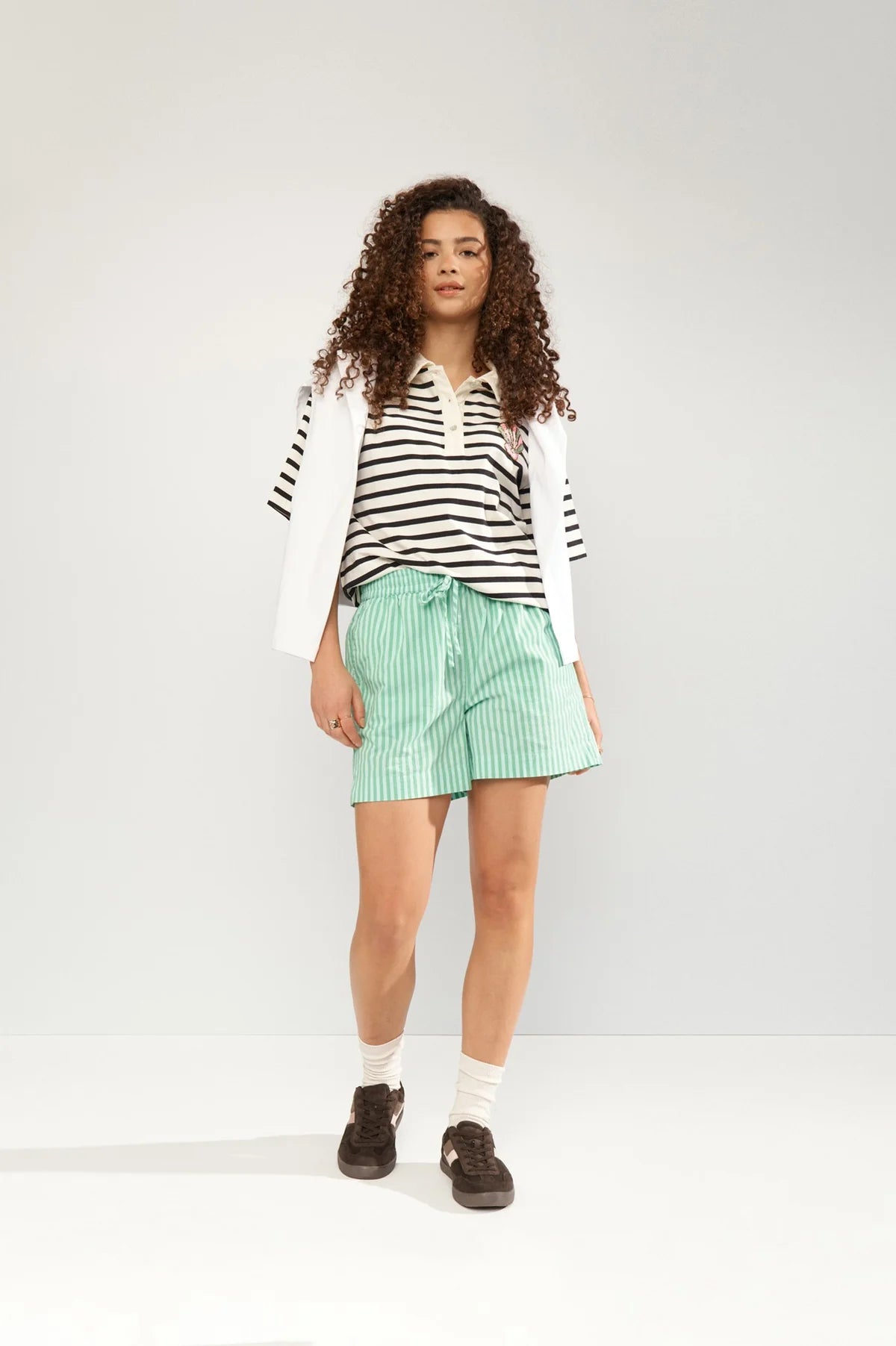 Person wearing a striped shirt, white jacket, green shorts, and black shoes on a white background