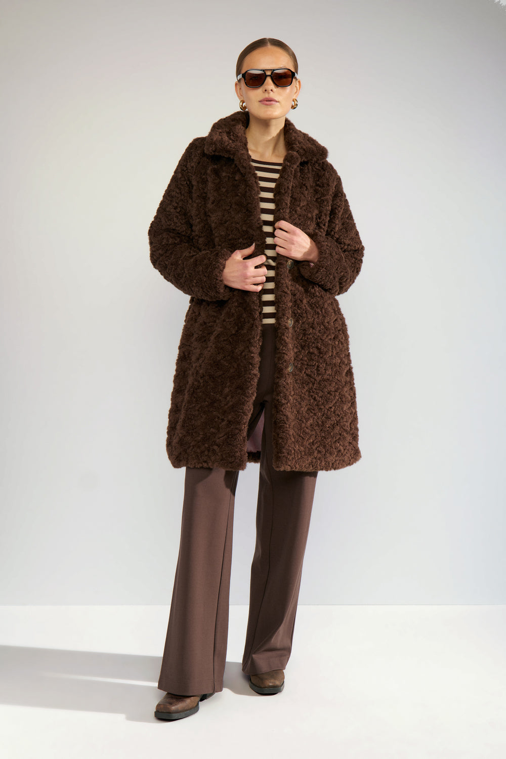 A model wearing a brown teddy coat, brown wide-leg trousers, a black and beige striped top, and sunglasses, standing against a light gray studio background.