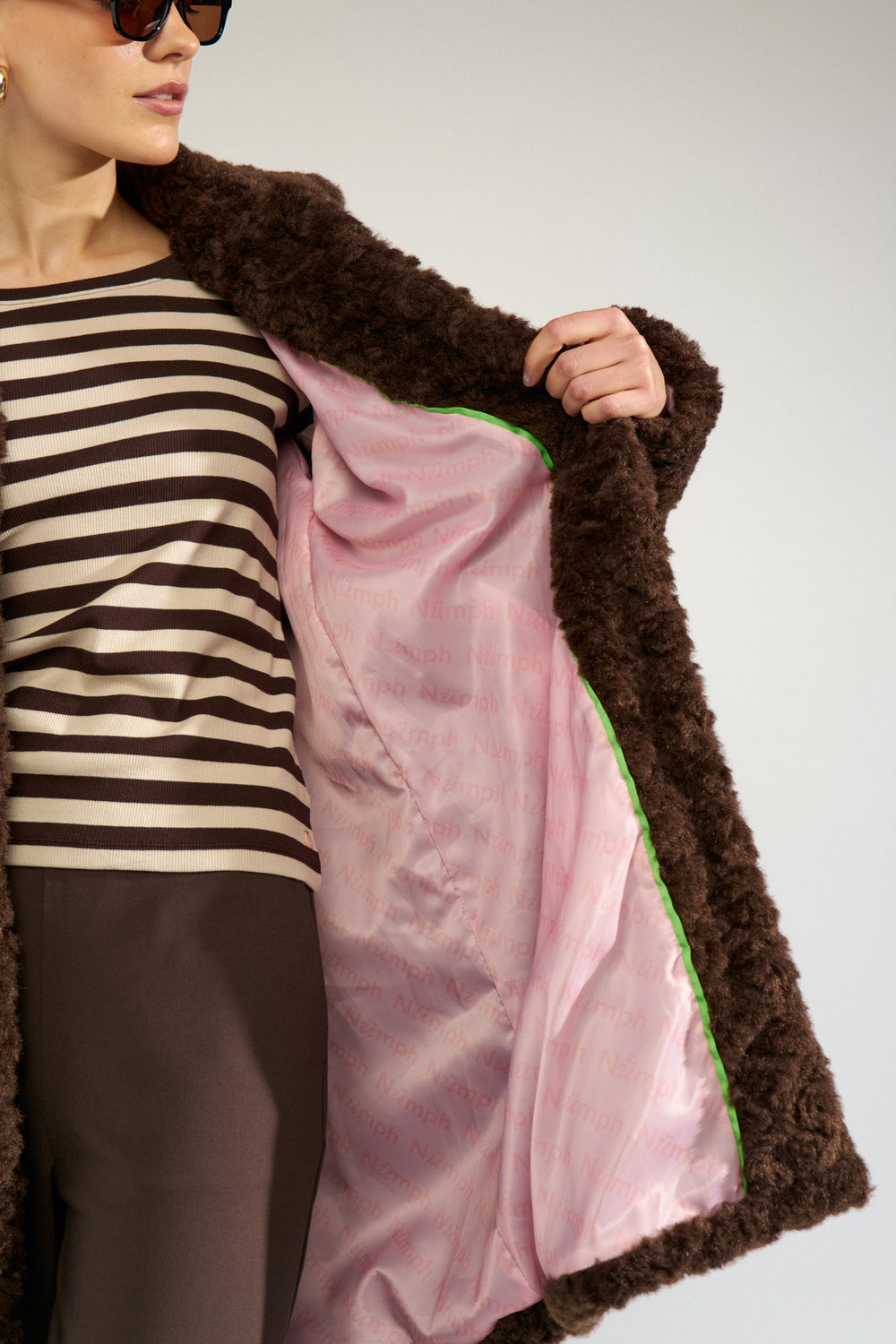 A close-up of a model wearing a dark brown curly faux-fur coat with a shiny light pink satin lining and a thin green trim, paired with a black and white striped top.
