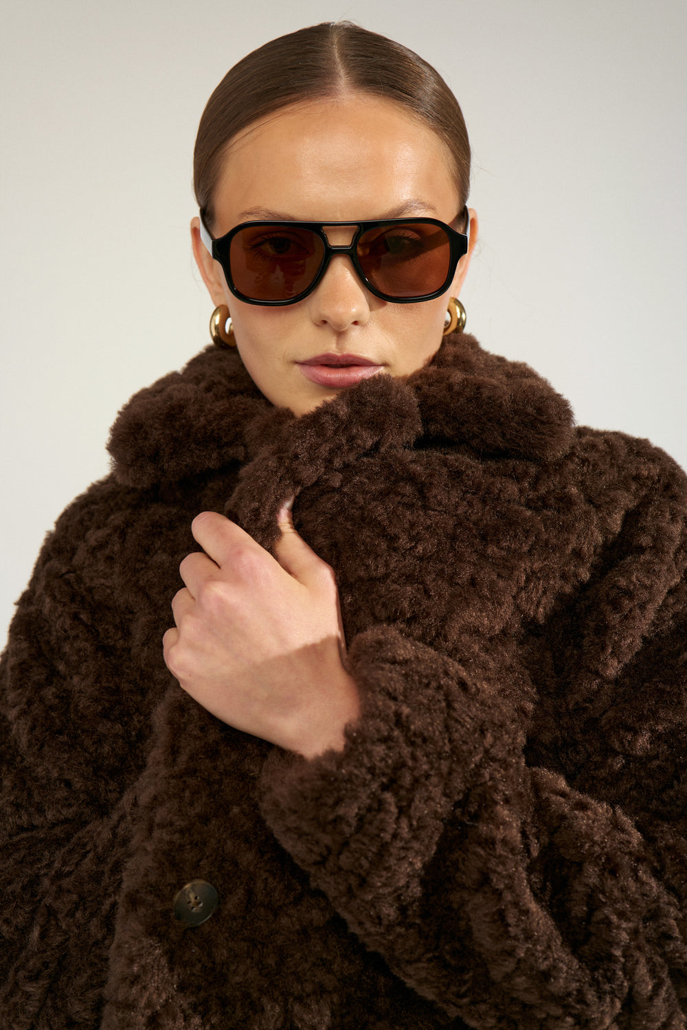 A model wearing a luxurious dark brown textured faux fur coat, black sunglasses, and gold hoop earrings, photographed against a neutral light gray background.