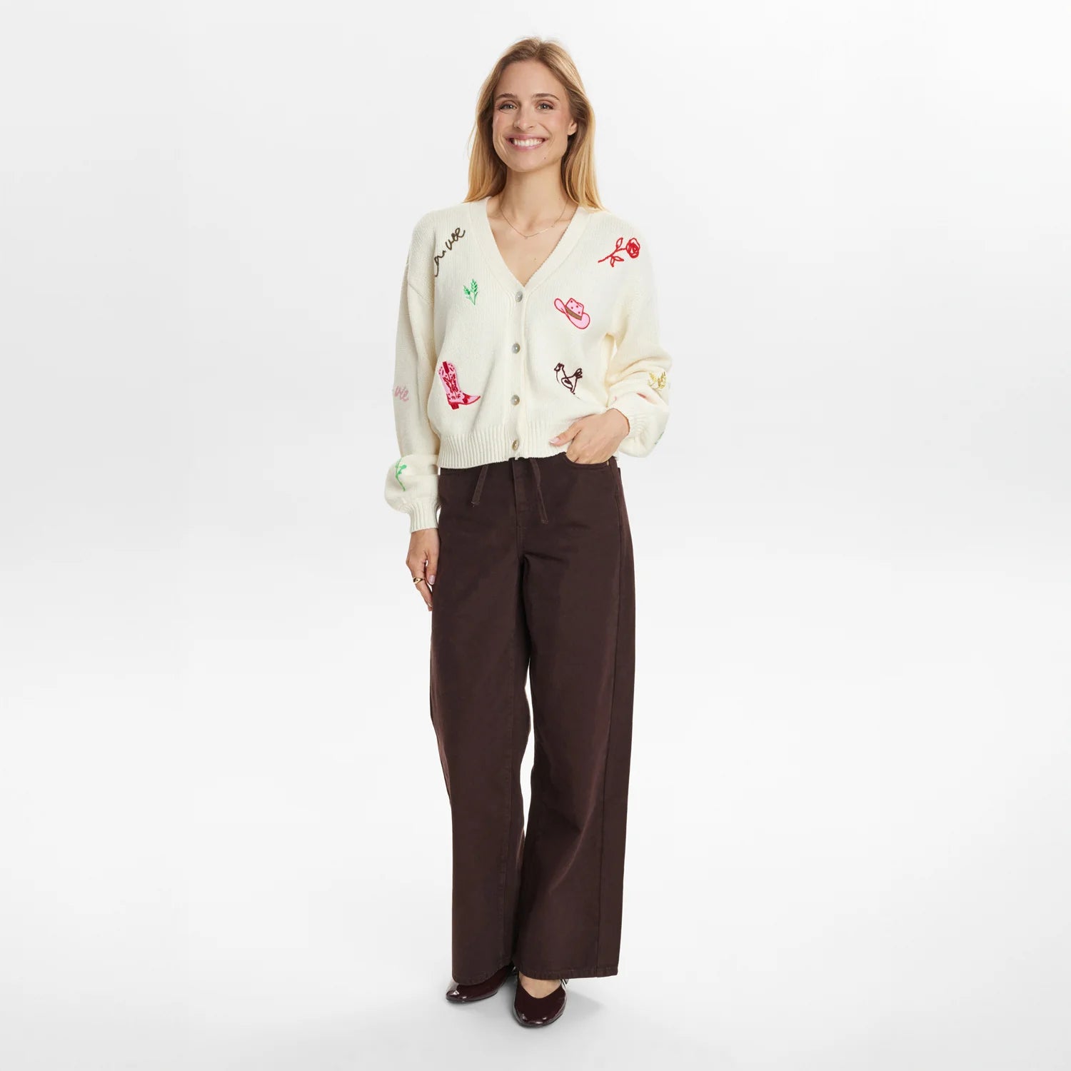 Woman wearing a cream cardigan with colorful embroidery and brown pants on a white background