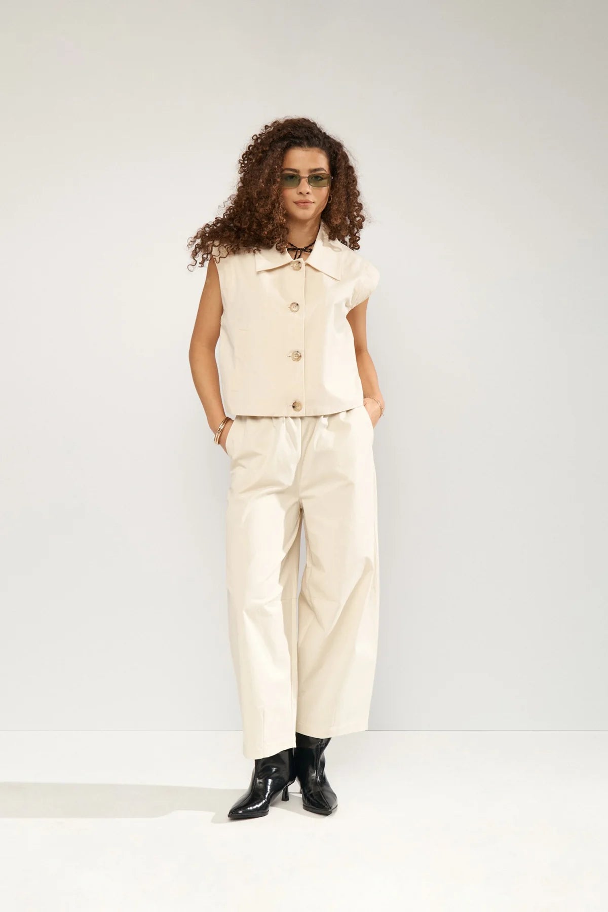 Woman wearing a cream sleeveless top and pants with black shoes on a white background