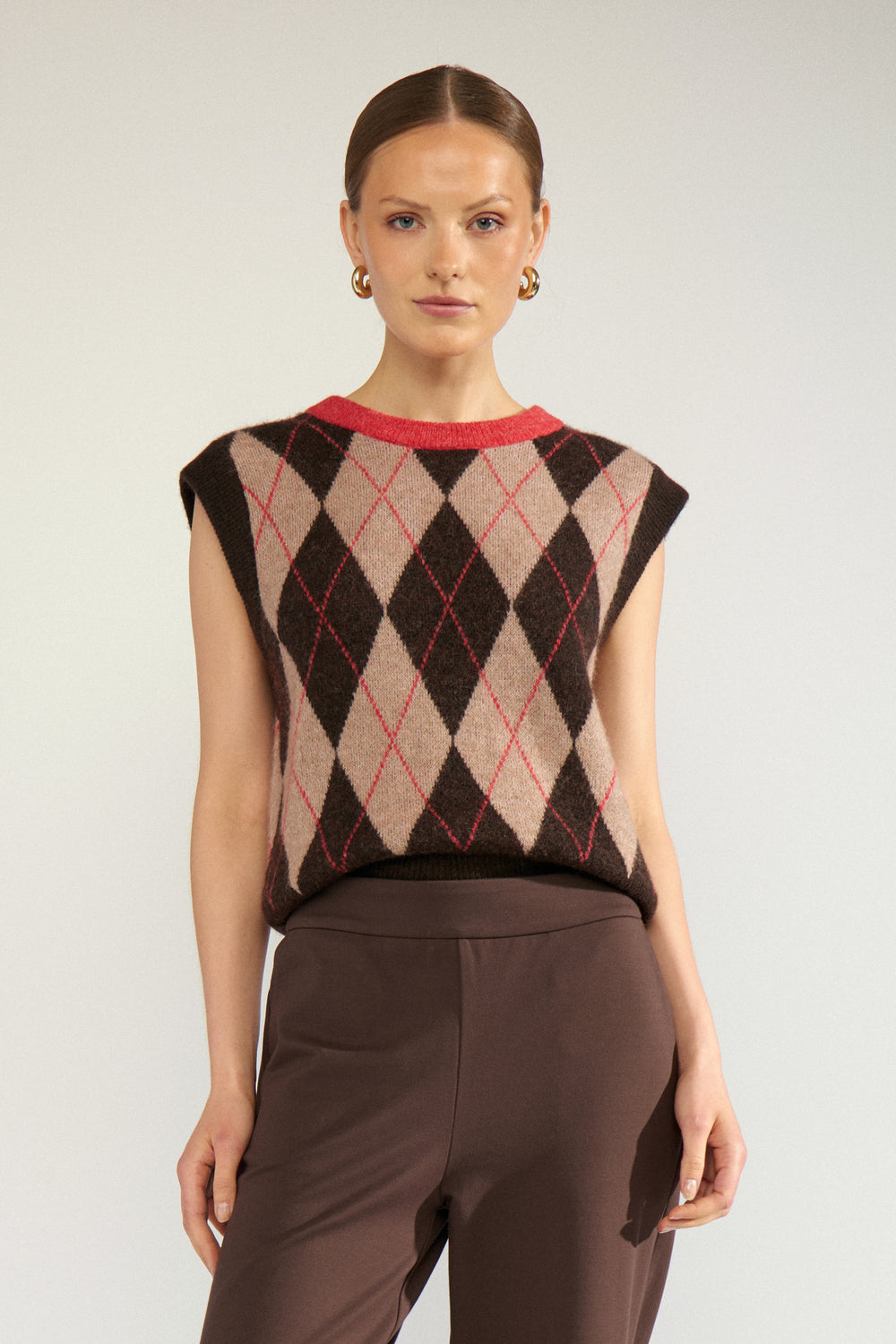 A woman modeling a sleeveless knit top with an argyle pattern in beige, brown, and red, paired with brown trousers, against a light gray studio background.