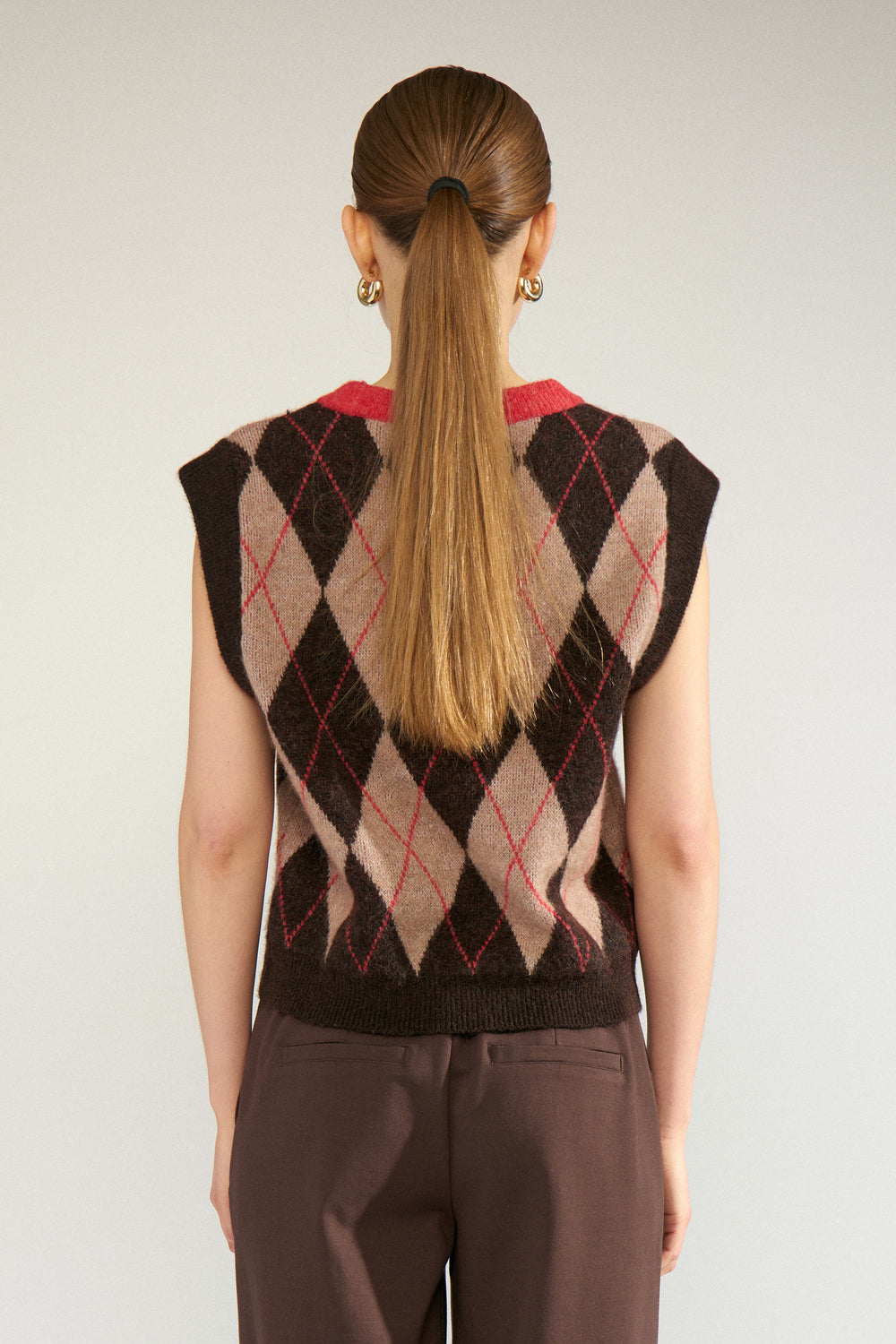 A woman viewed from the back wearing a sleeveless argyle sweater vest with brown, beige, and red diamonds, paired with brown trousers, against a light gray studio background.