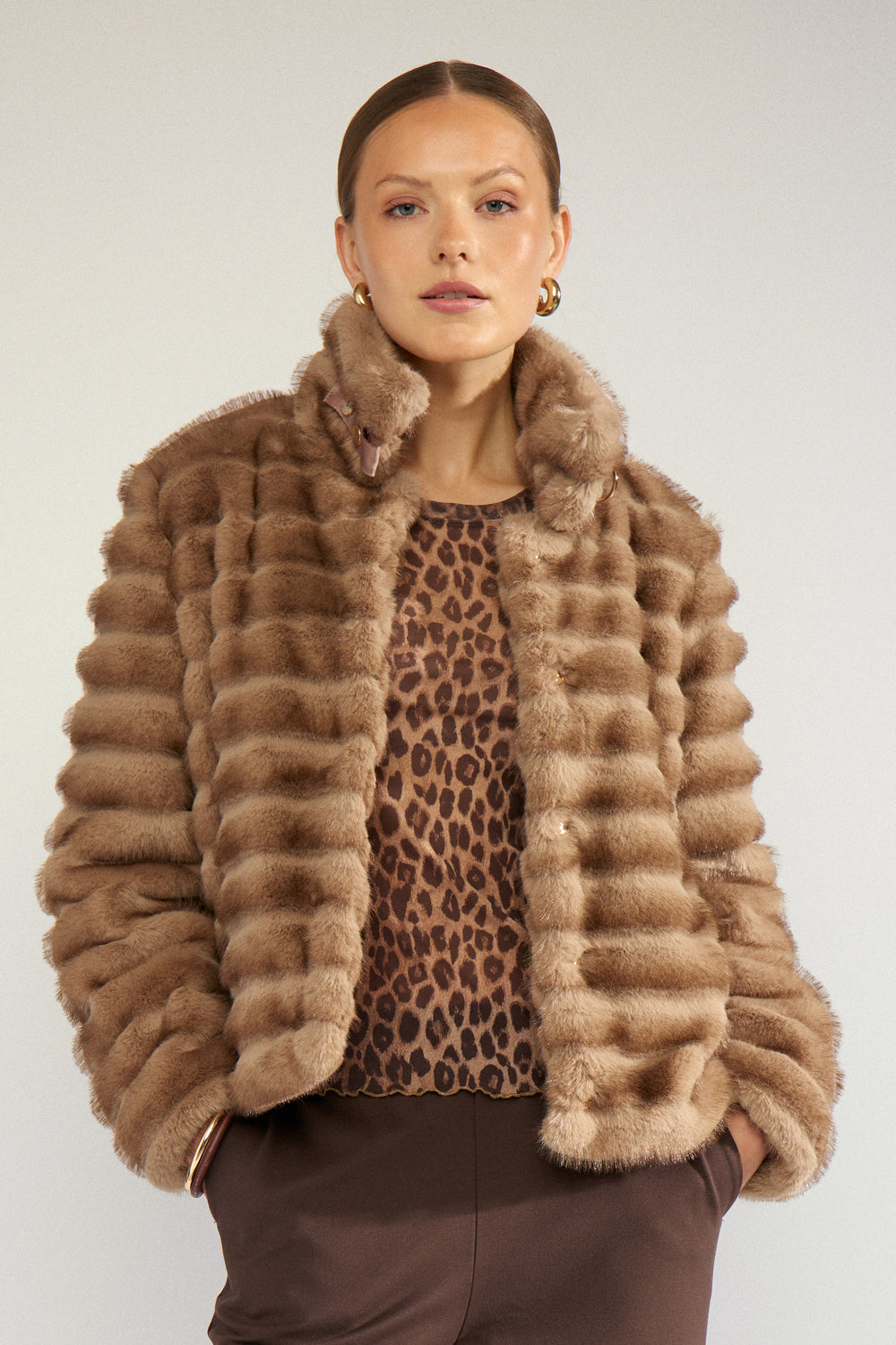 A model wearing a brown and beige striped fur jacket over a leopard-print top and brown trousers, photographed against a light gray studio background.
