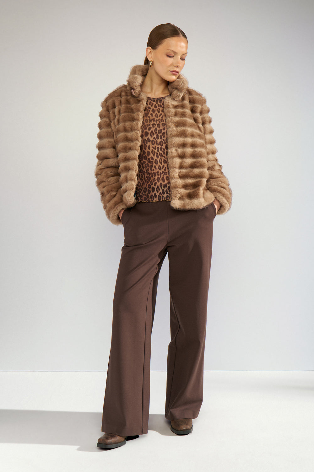 A model stands against a light gray studio background wearing a brown shaggy faux-fur jacket over a leopard-print top and dark brown wide-leg trousers.