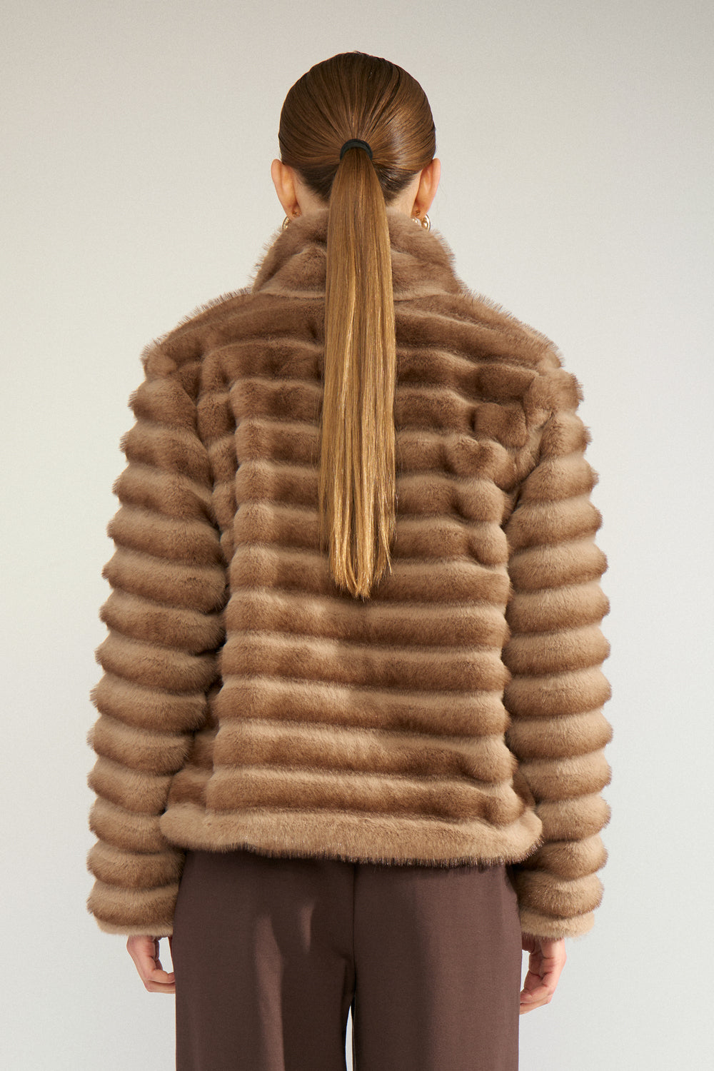 A person shown from the back wearing a brown fur jacket with horizontal stacked texture and dark brown pants, photographed against a plain light gray studio background.
