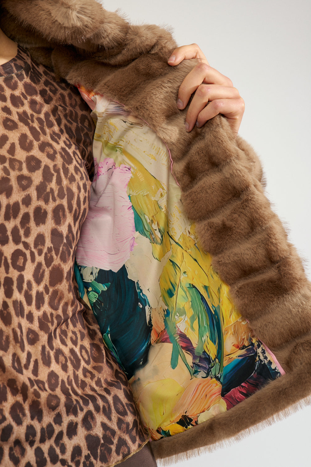A close-up of a person wearing a brown fur-trimmed jacket over a leopard-print top, with a colorful abstract-patterned scarf or fabric draped across the chest against a light gray background.