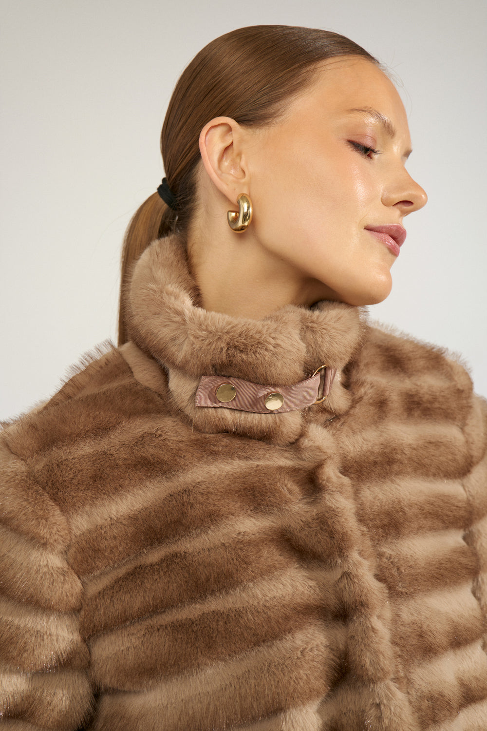 A close-up side profile of a woman wearing a brown and beige striped fur jacket with a high padded collar and gold hoop earrings, against a light gray background.