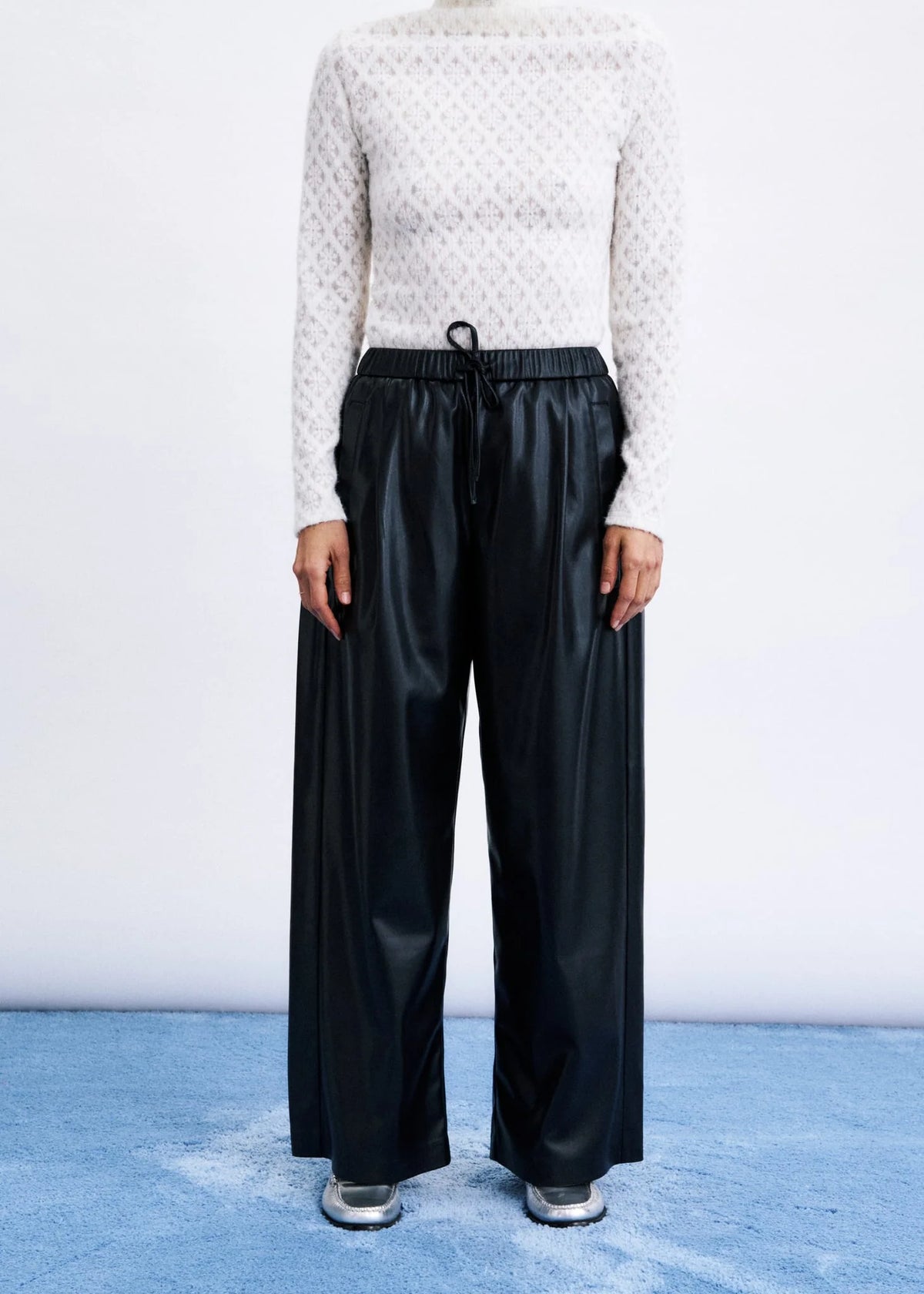 A model is shown from the neck down wearing black wide-leg faux-leather trousers with a drawstring waist, paired with a white textured knit sweater and silver shoes, standing on a blue textured floor against a white studio background.