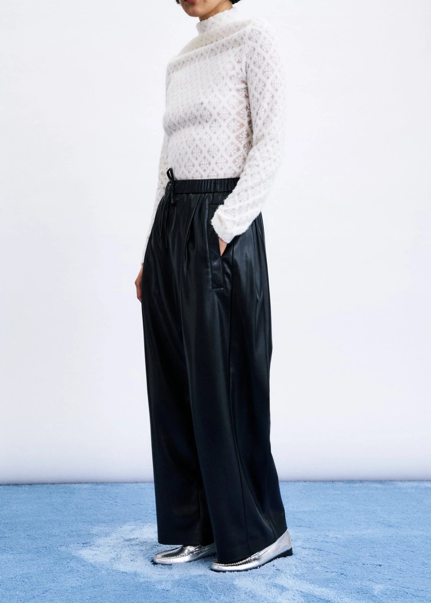 A model wearing a white textured knit mock-neck sweater and black wide-leg trousers, standing against a white studio wall on a blue floor.