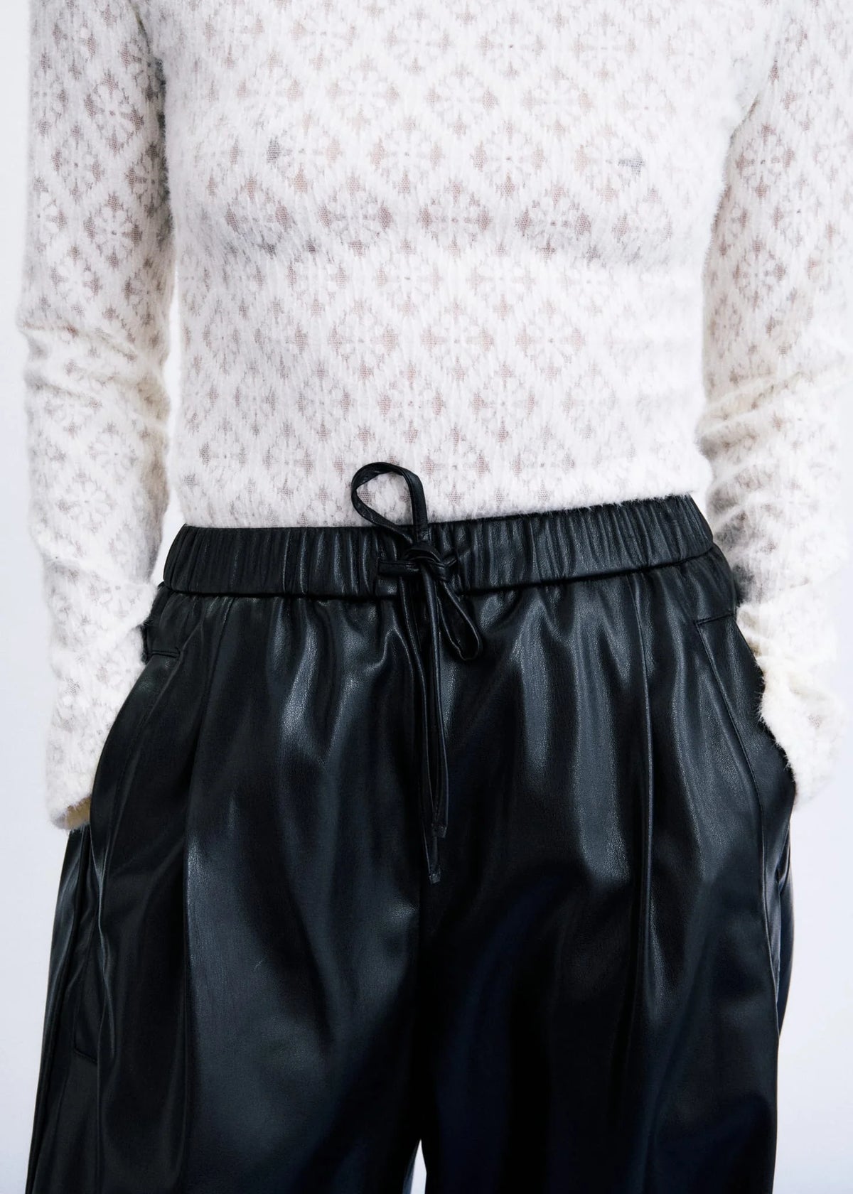 A close-up of a person wearing black faux-leather drawstring trousers and a white textured knit sweater against a plain light background.