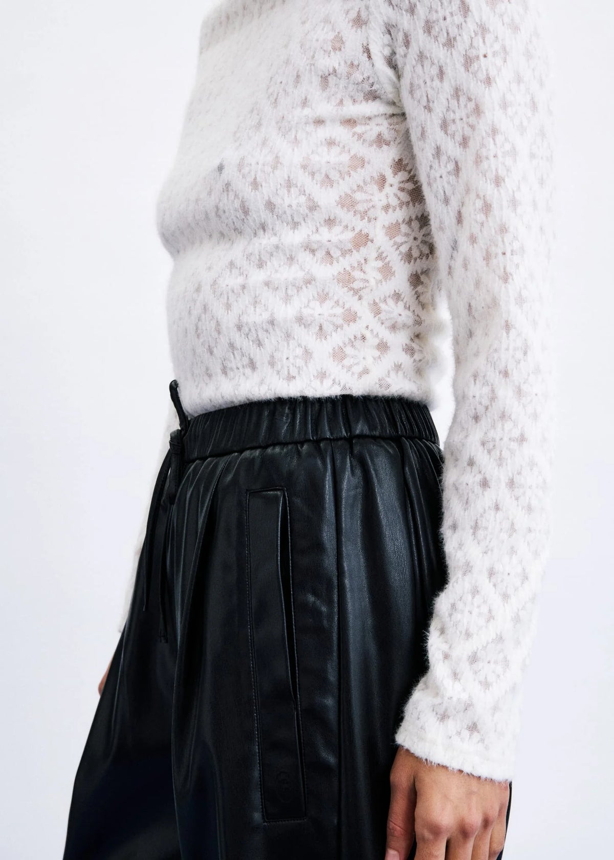 A close-up of a person wearing a white textured knit sweater and black faux-leather trousers against a plain light background.