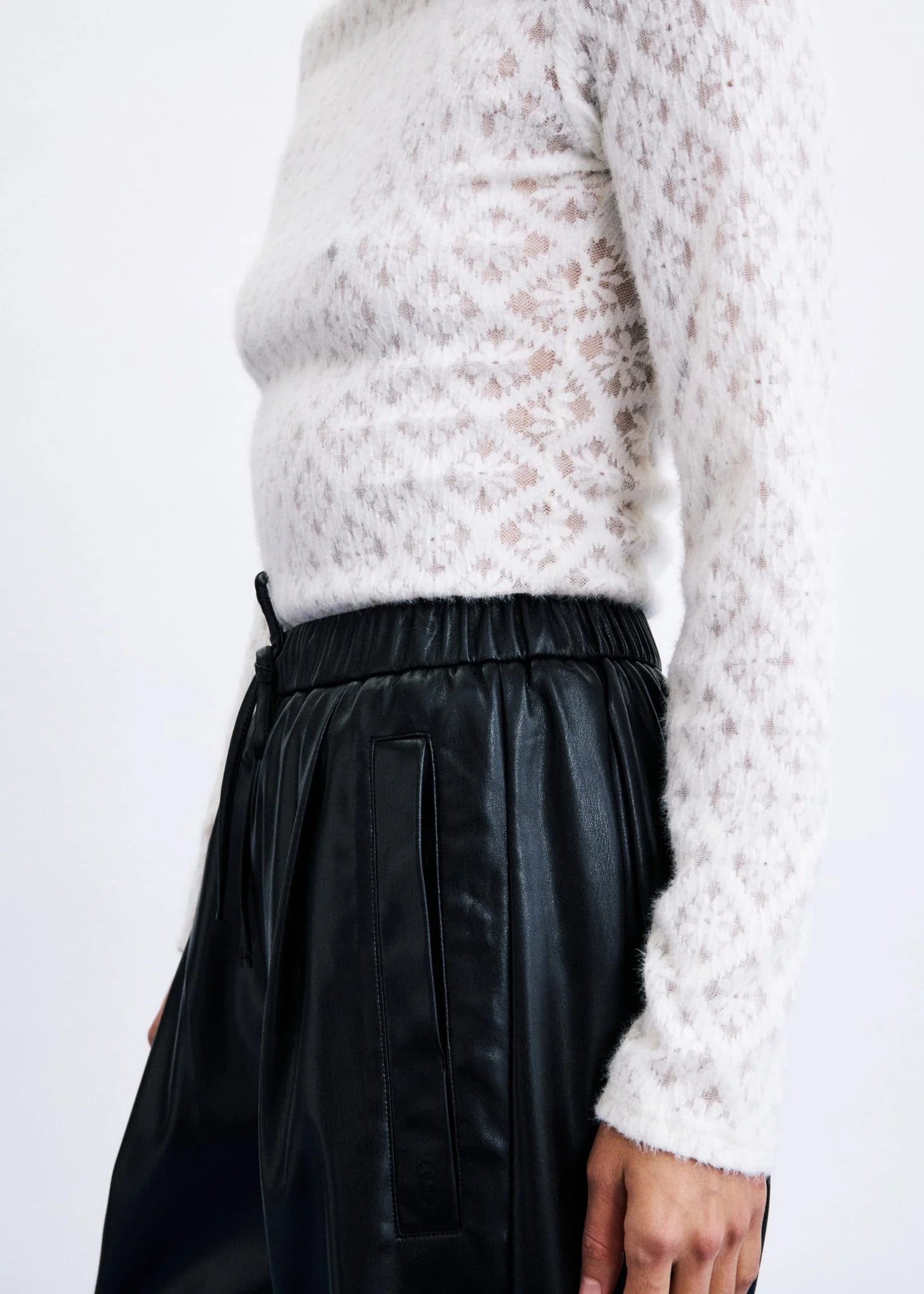 A close-up of a person wearing a white textured knit sweater and black faux-leather trousers against a plain light background.