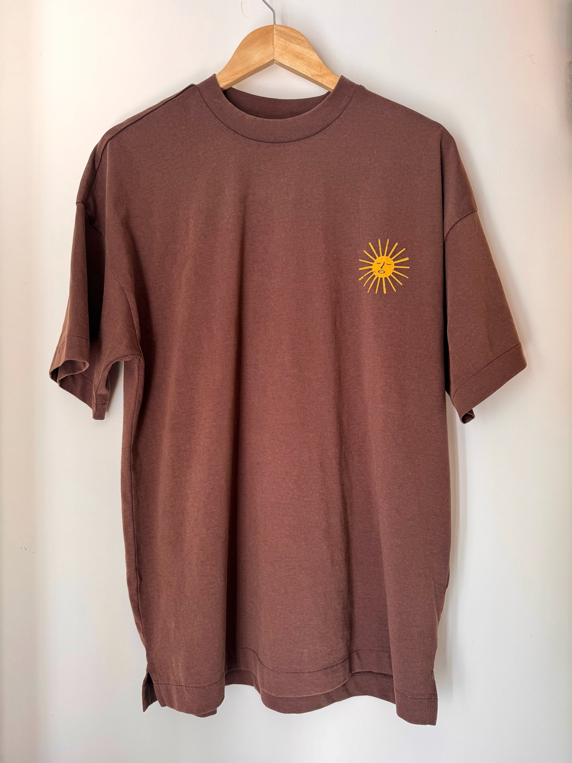Brown t-shirt with a yellow sun design on a hanger against a white wall