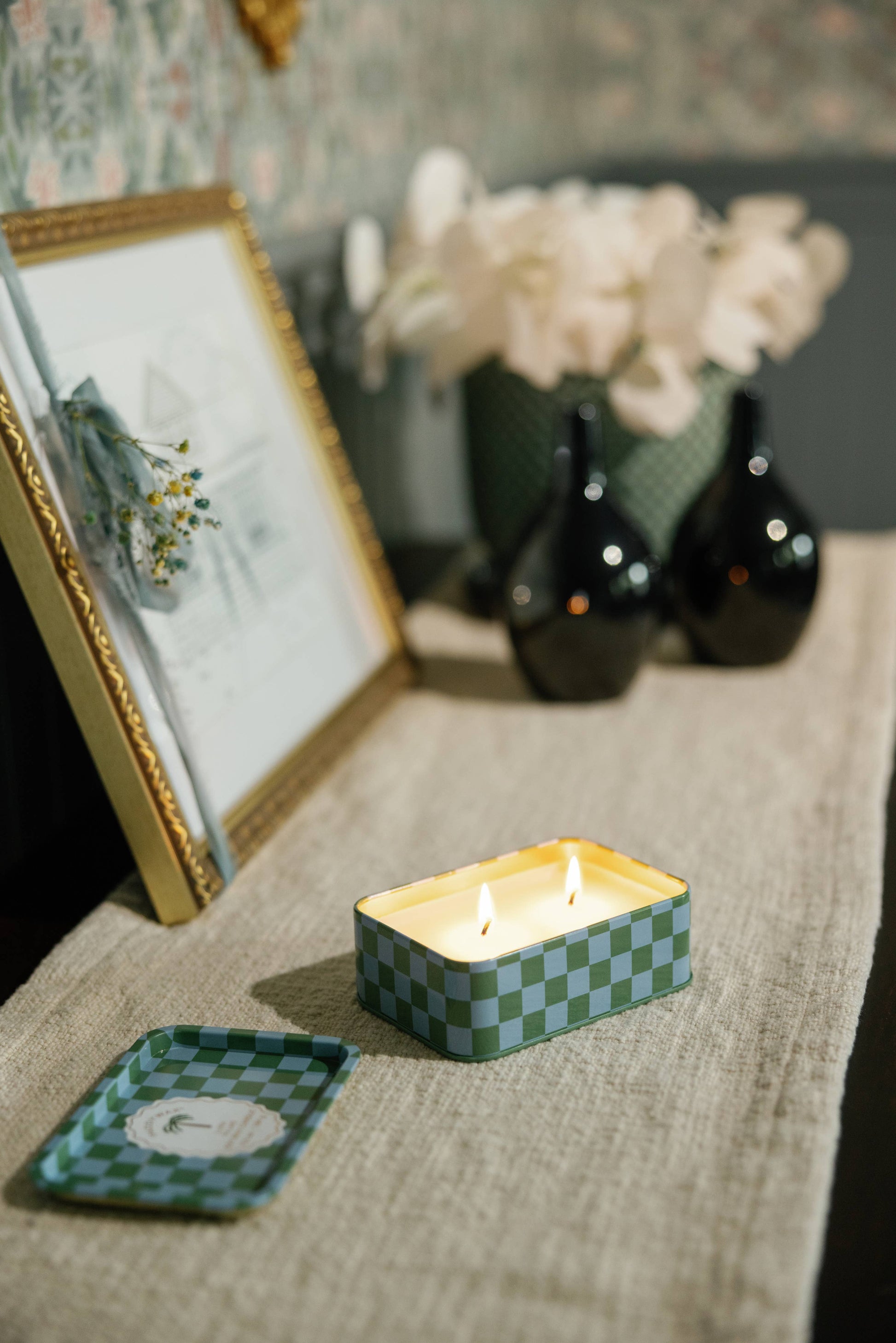 Candle in a checkered tin on a textured surface with a framed picture and flowers in the background.