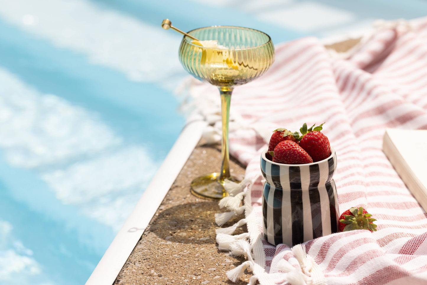Cocktail glass with a straw, strawberries in a striped container, and a book on a towel by a pool.