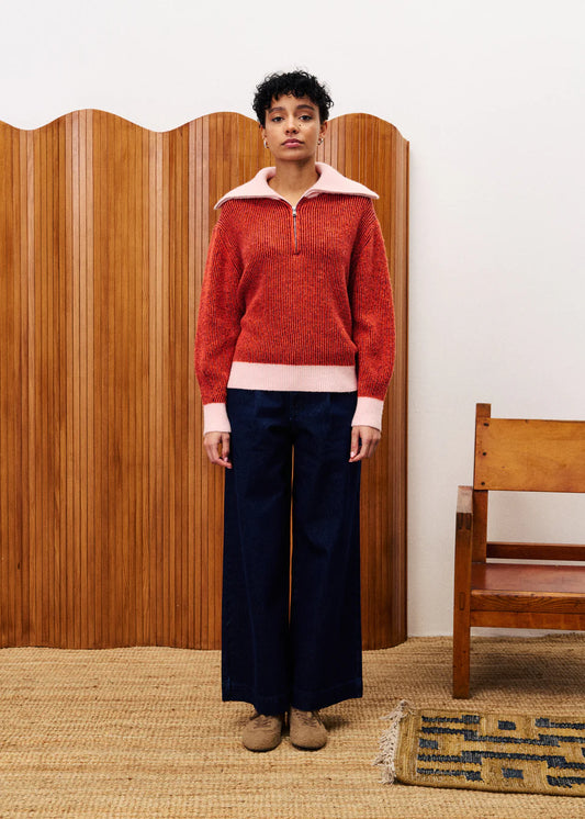 Person wearing a red sweater with white collar and navy pants standing in front of a wooden screen.