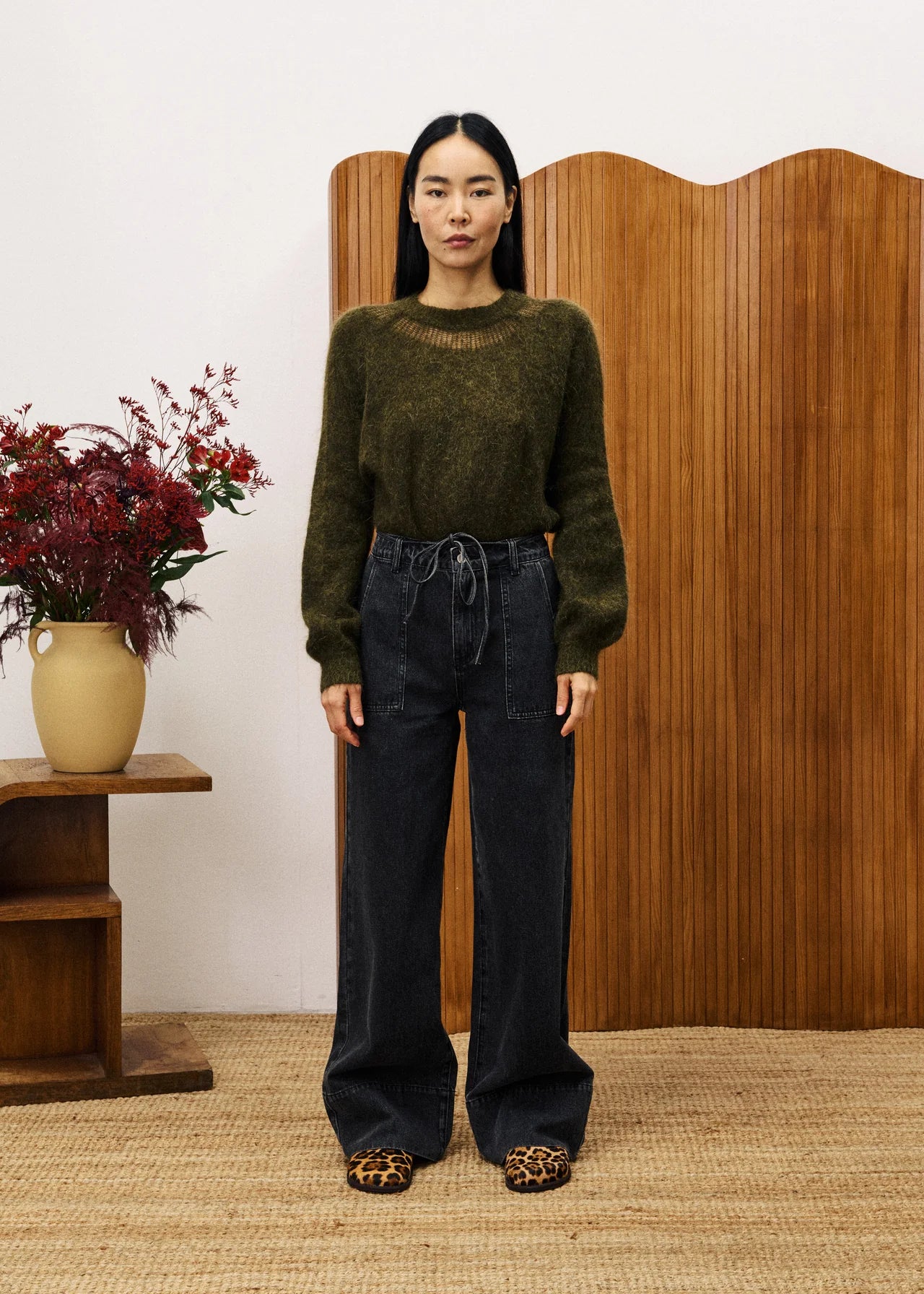 Woman wearing a green sweater and black pants standing in front of a wooden screen with a vase of flowers on a table.