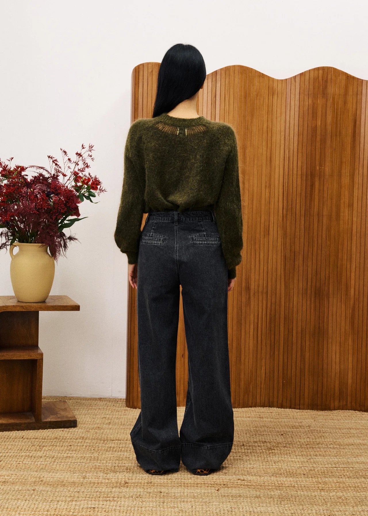 Person wearing a green sweater and dark jeans standing in front of a wooden screen with a vase of flowers on a table.