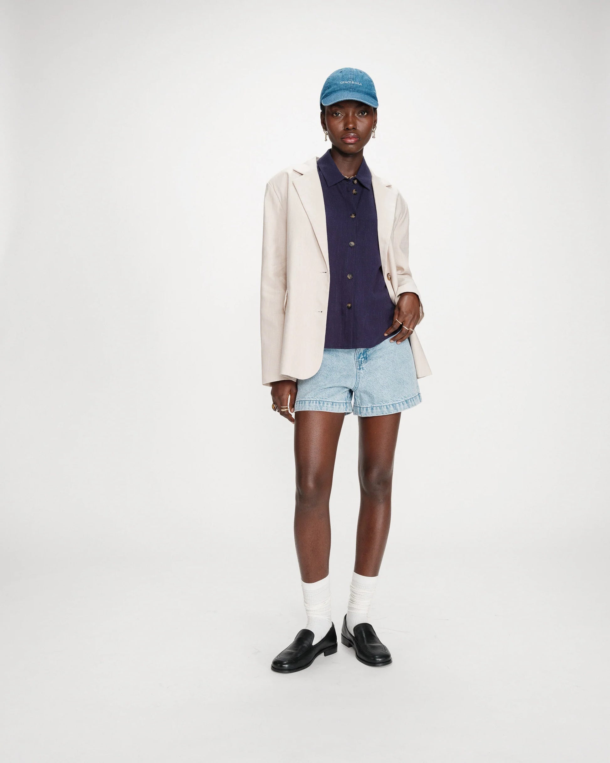 Person wearing a beige blazer, navy shirt, denim shorts, and black shoes on a white background