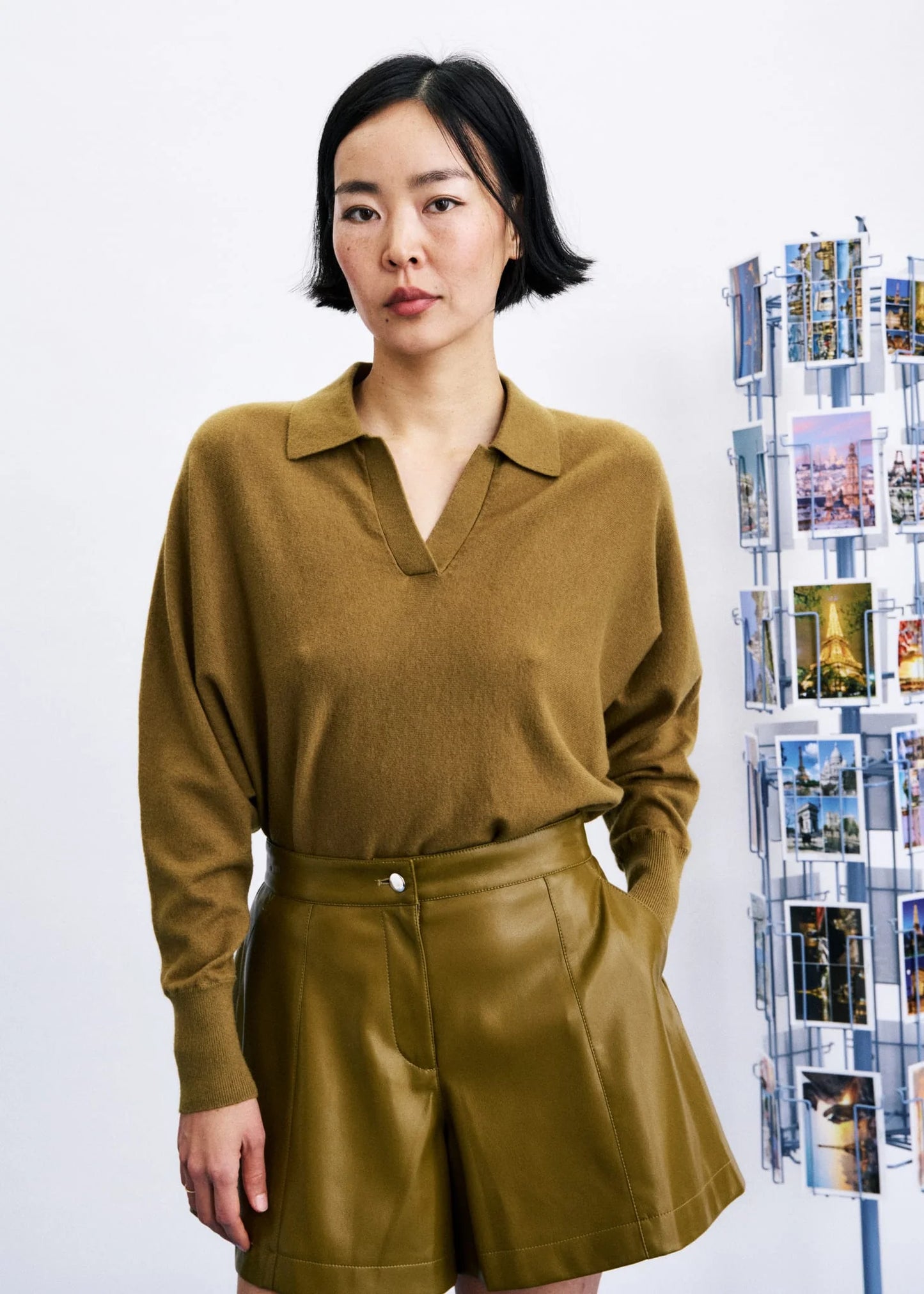 A woman wearing a camel brown knit polo shirt and matching brown shorts, standing against a white wall with a photo collage board on the side.
