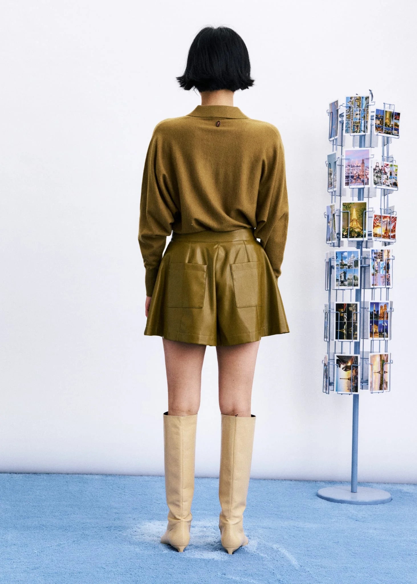 A model viewed from behind wearing a matching olive green long-sleeve knit top and faux-leather mini skirt, paired with beige knee-high boots, standing on a blue carpet next to a photo display stand.