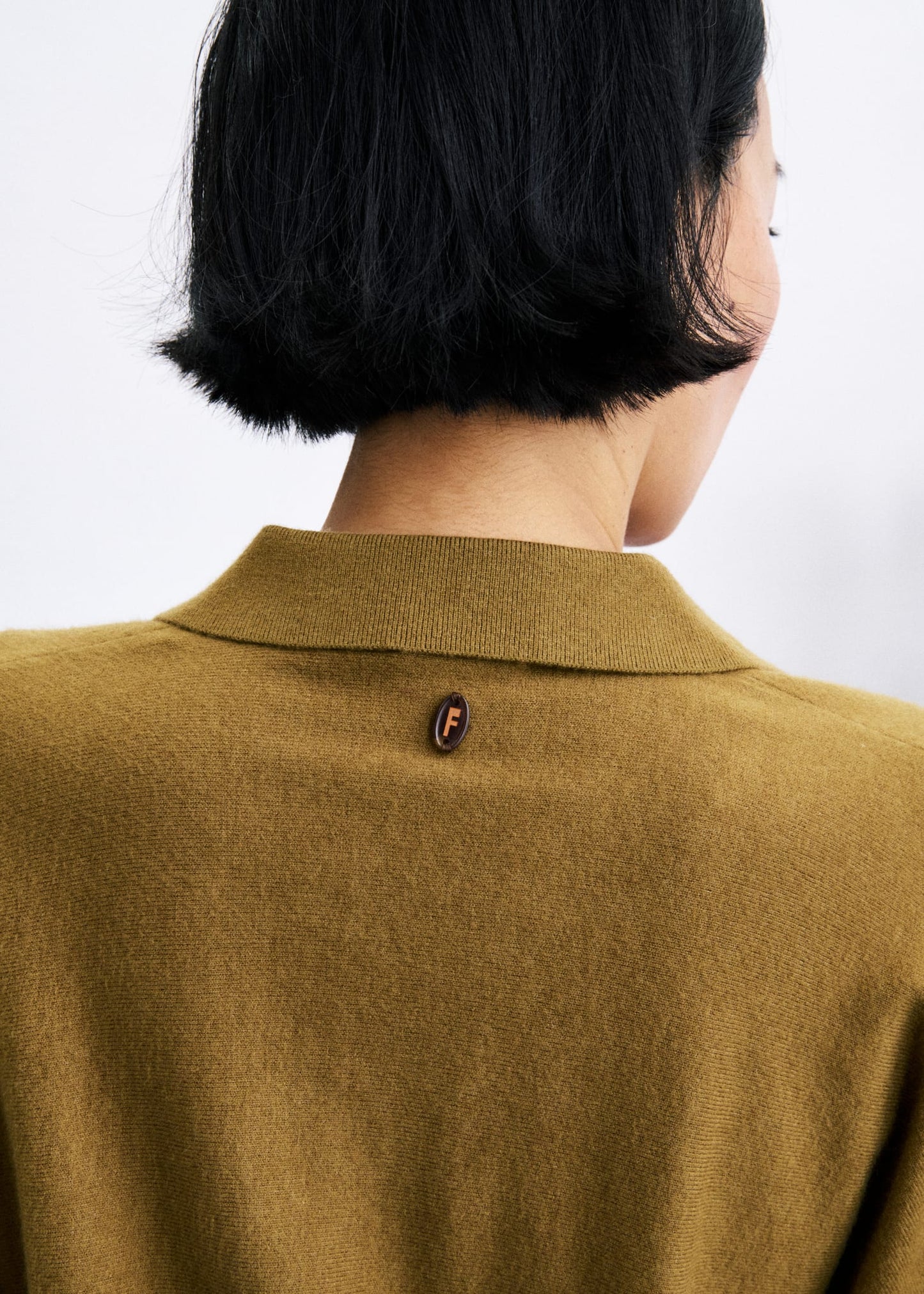 Close-up back view of a person wearing a camel-colored knit polo shirt with a small embroidered Fendi logo on the collar.