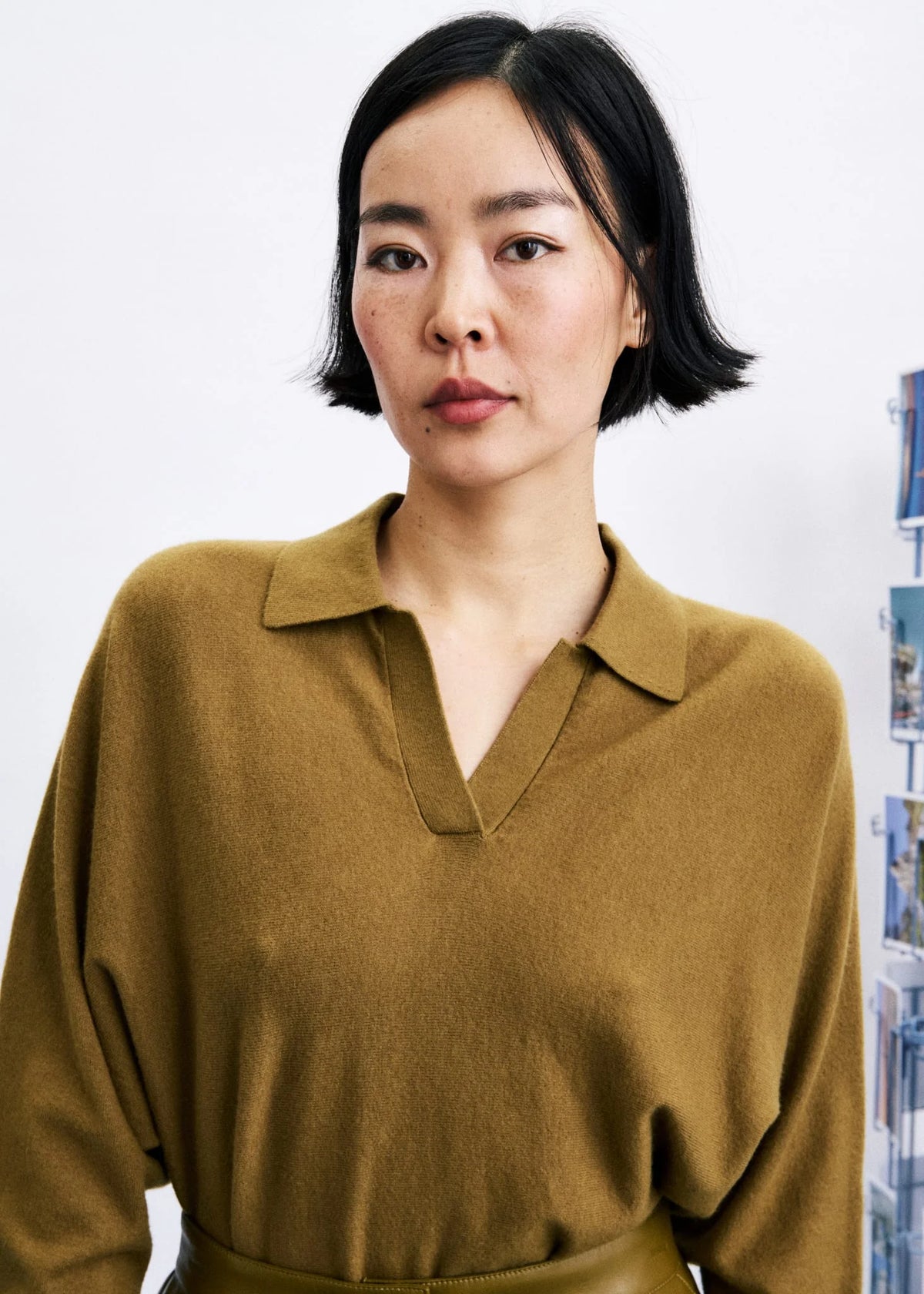 A woman with short black hair wearing a mustard yellow knit polo shirt, shown from the chest up against a light gray wall.